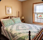 Small bedroom with a wooden four-poster bed covered in a green-and-white quilt, a nightstand with a lamp, framed artwork on the wall, and a window with a blind and small stained-glass decoration.