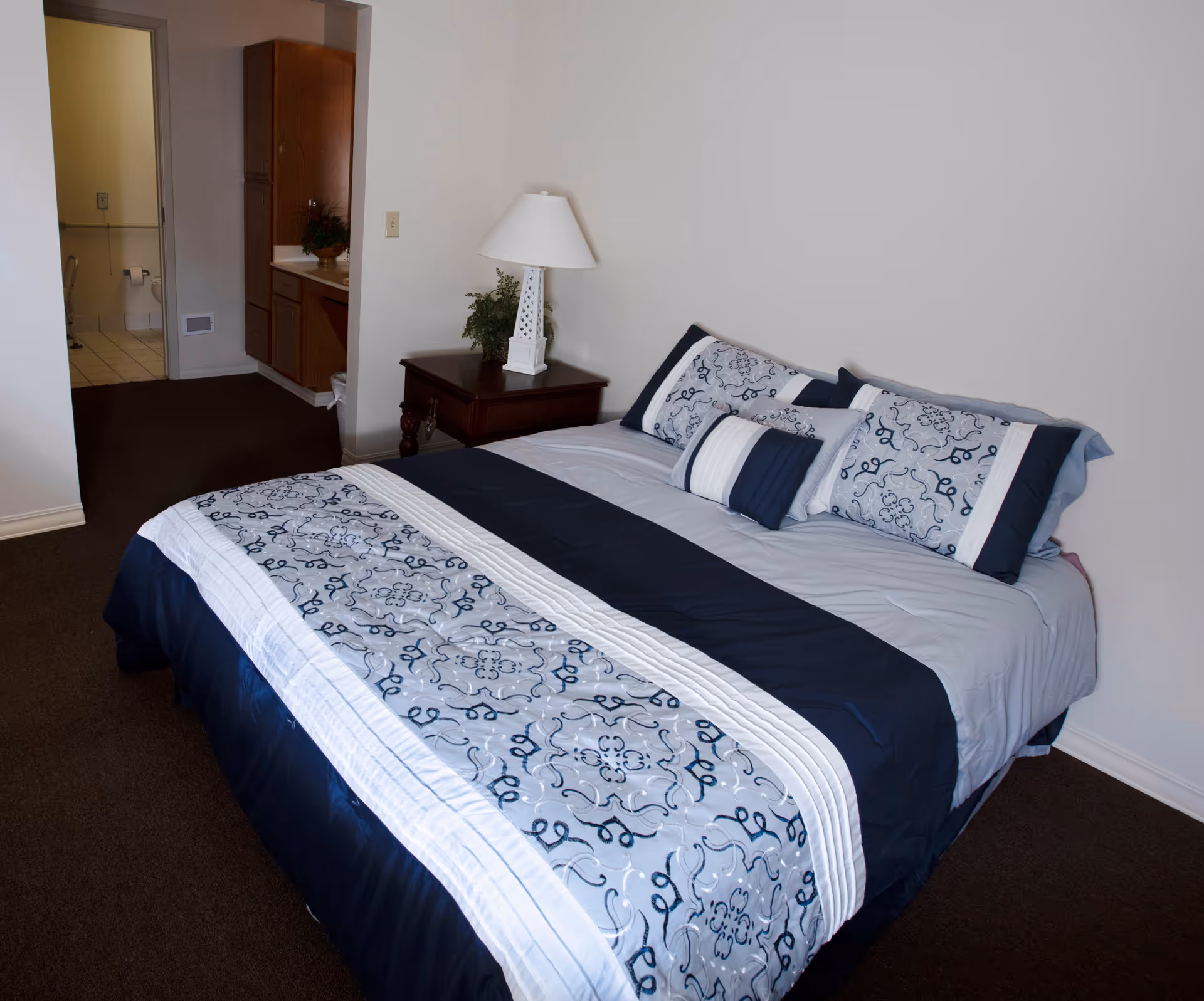 Neatly made bed with blue-and-white patterned bedding in a simple bedroom with a nightstand, lamp, and an open doorway to a bathroom.
