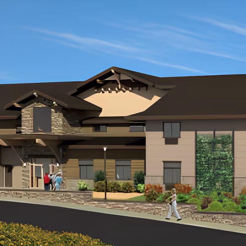 Exterior view of a senior living facility building with a stone and beige facade, large windows, and a landscaped garden area. A person is walking on the sidewalk in front of the building under a clear blue sky.