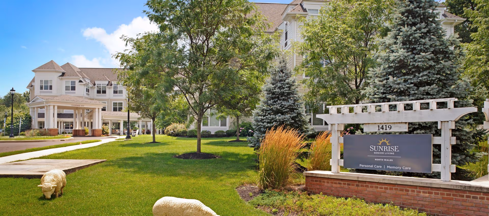 Exterior view of Sunrise Senior Living facility in North Wales, showing a large, multi-story building surrounded by green trees and well-maintained lawn with decorative sheep statues. A sign in front reads 'Sunrise Senior Living North Wales Personal Care | Memory Care'.