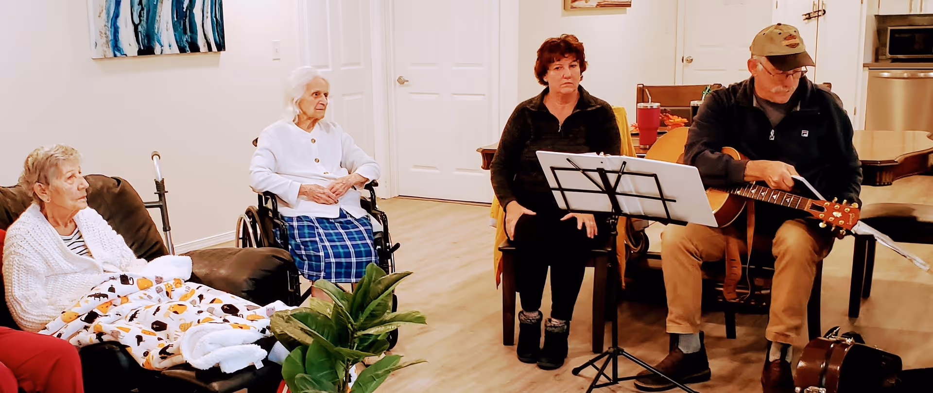 A group of elderly people and a caregiver in a living room setting. Two elderly women are seated, one in a wheelchair and the other on a recliner with a blanket. A woman and a man with a guitar are seated nearby, with the man preparing to play music from a stand with sheet music. The room has light-colored walls and wooden flooring.
