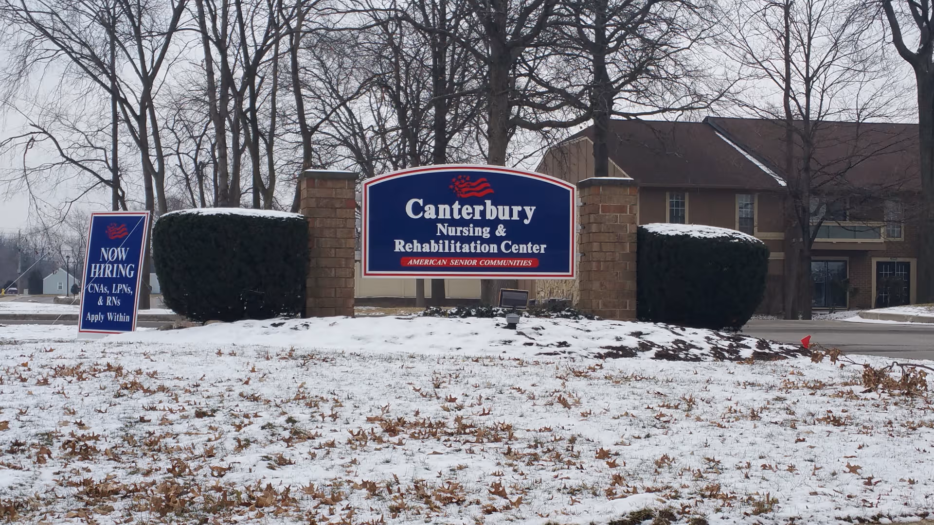 Canterbury Nursing and Rehabilitation Center entrance sign with a Now Hiring board in snow.