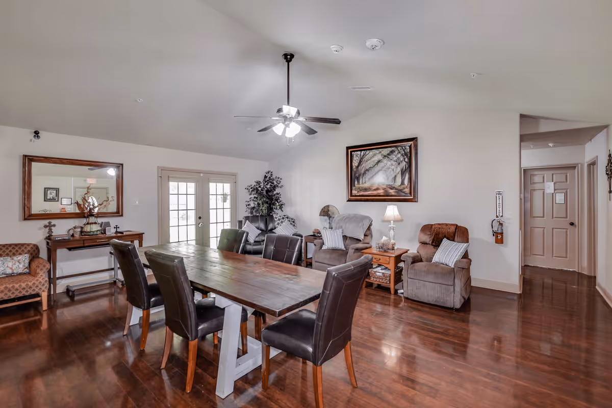 A cozy common area with a wooden dining table surrounded by six black leather chairs. The room features a ceiling fan with lights, a large mirror on the wall above a console table with decorative items, and several comfortable armchairs with pillows and blankets. A framed picture of a tree-lined path hangs on the wall, and double glass doors lead outside. The floor is polished wood, and the walls are painted light beige.
