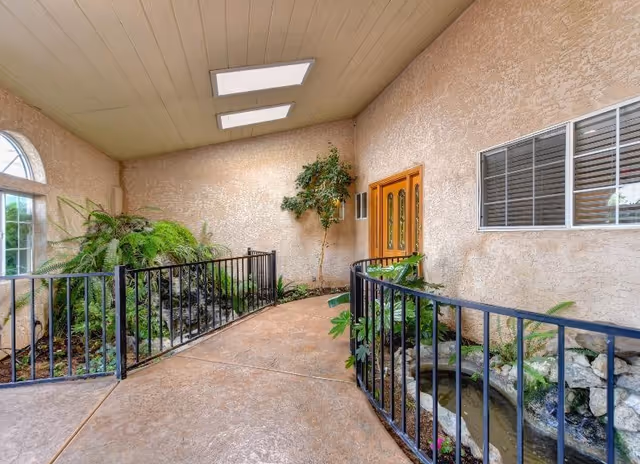 Covered entrance with a sloped concrete ramp flanked by black railings, plants and a small water feature leading to double wooden doors.