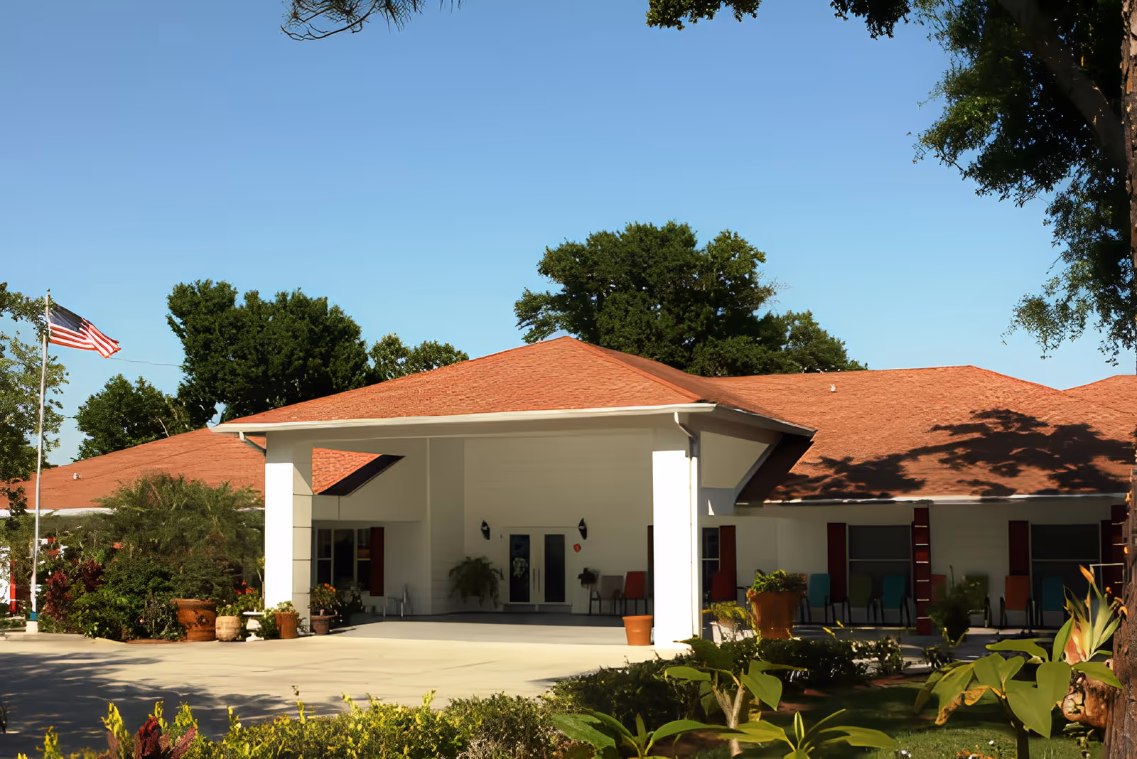 Front exterior of a single-story senior living building with a covered entrance, landscaping, and an American flag.