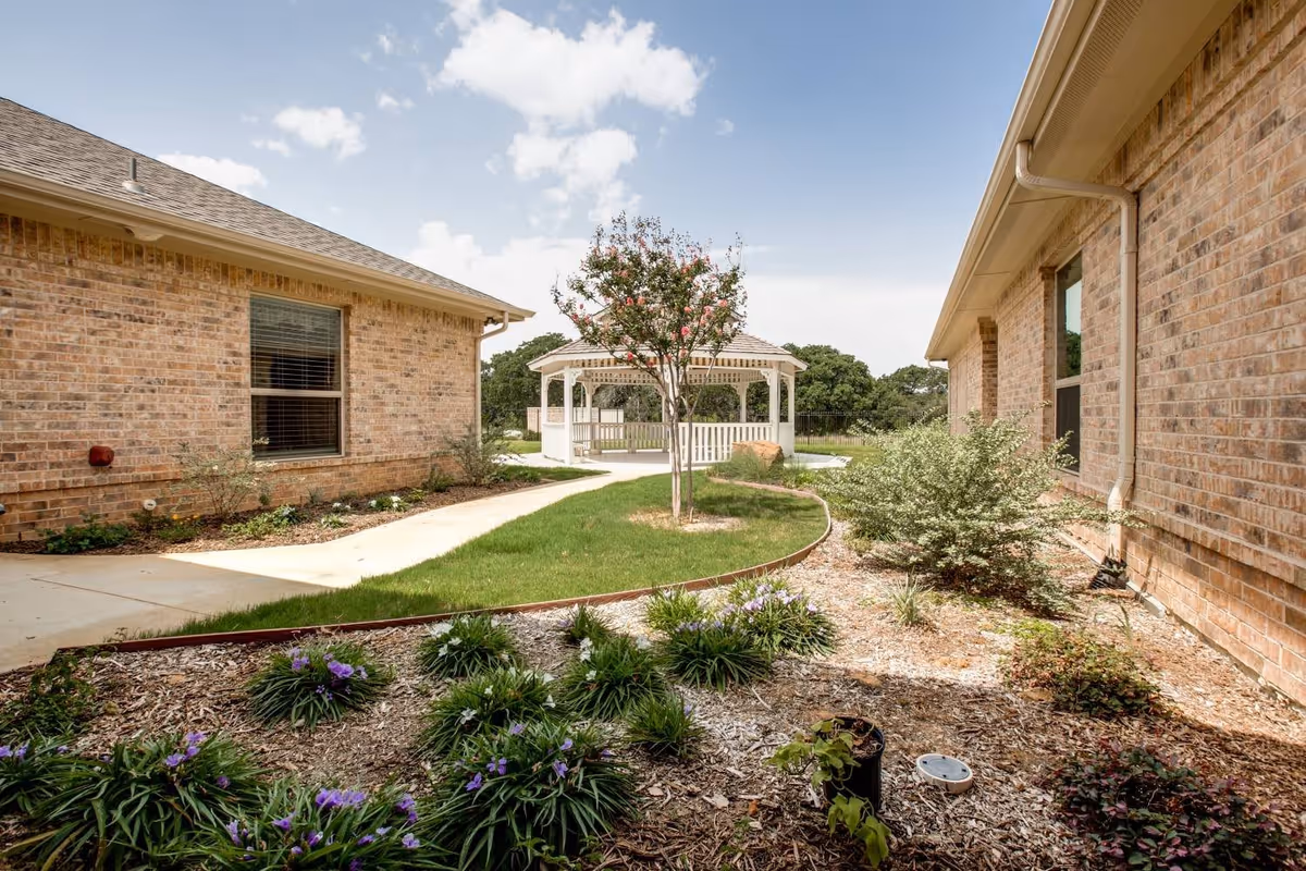 Outdoor garden area between two brick buildings with a small tree, various plants, and a white gazebo in the background under a partly cloudy sky.