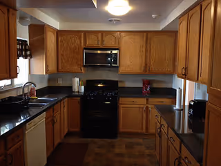 A kitchen with wooden cabinets, black countertops, a black stove with a microwave above it, a dishwasher, a sink with a faucet, and various kitchen items such as a red coffee maker and paper towels.