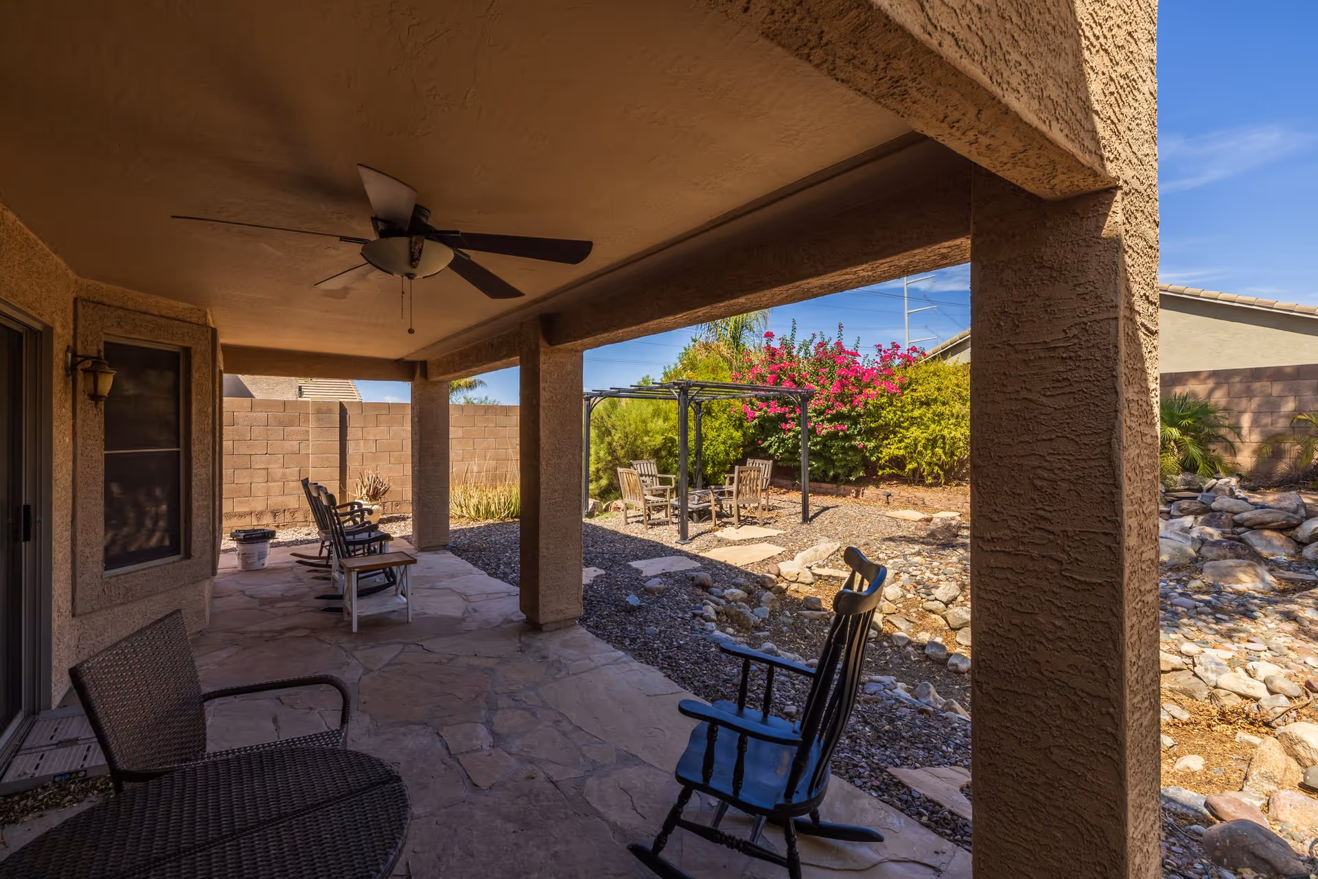 Covered patio area with ceiling fan and several chairs, overlooking a landscaped backyard with a small seating area under a pergola, surrounded by bushes and a stone pathway.