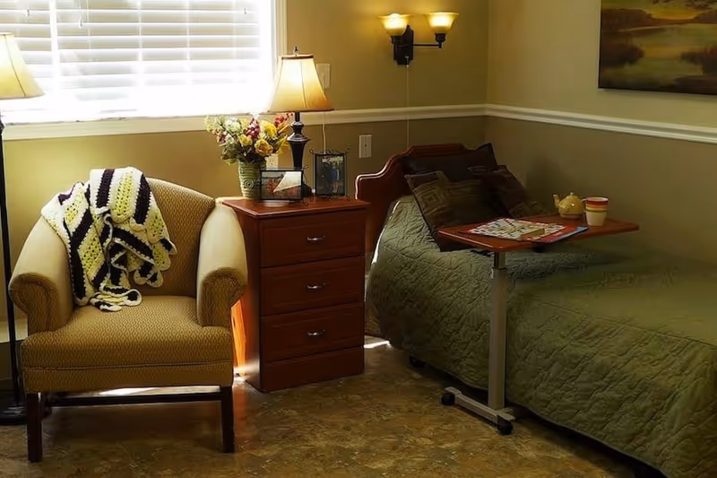A cozy bedroom with an armchair draped with a blanket, a nightstand holding a lamp and flowers, and a bed with a green quilt and an overbed table.