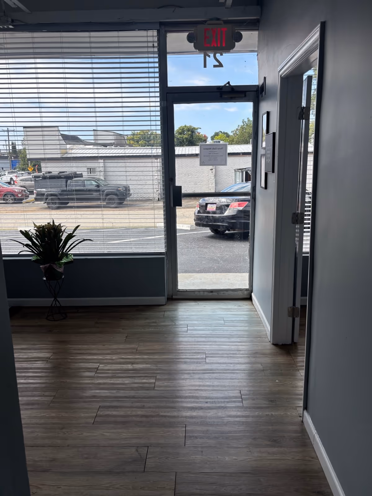 Indoor hallway with glass door, blinds, potted plant, and outdoor parking visible.