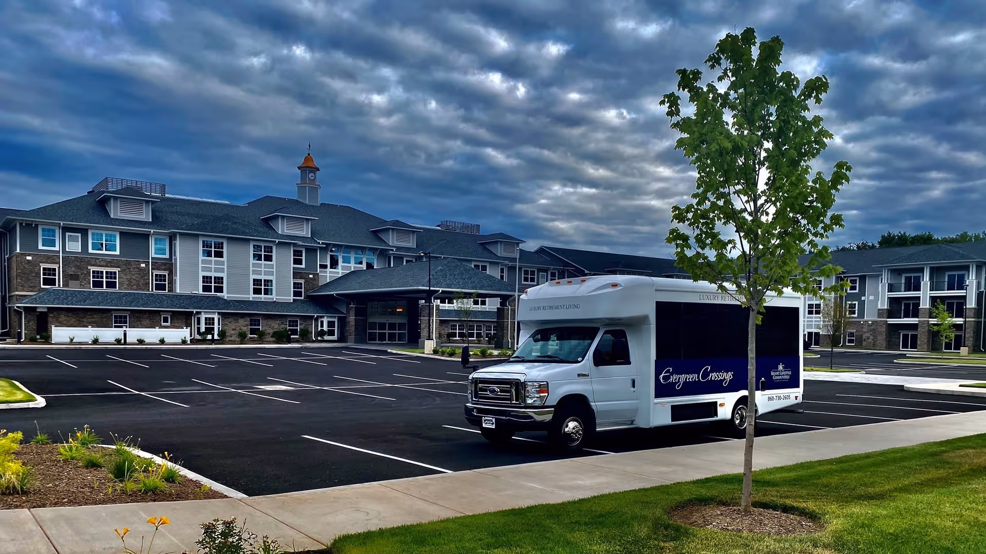 A large multi-story retirement community building with a clock tower on the roof under a cloudy sky. In the foreground, there is a mostly empty parking lot with a white shuttle bus parked near a young tree. The shuttle bus has signage that reads 'Evergreen Crossings'.