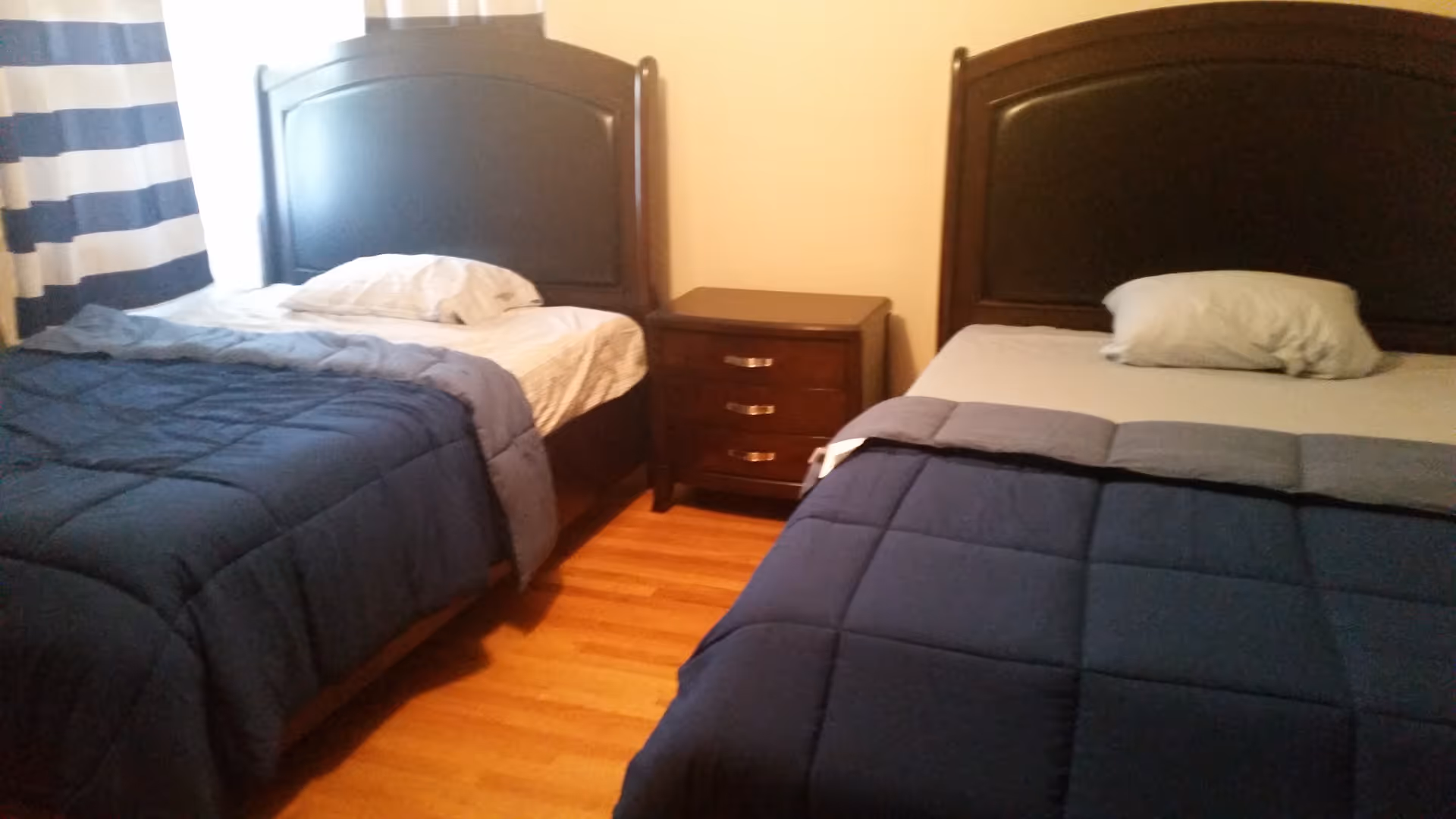A bedroom with two single beds, each with a dark wooden headboard. Both beds have blue comforters and white pillows. There is a small wooden nightstand with three drawers placed between the beds. The floor is wooden, and a window with blue and white striped curtains is partially visible on the left side.