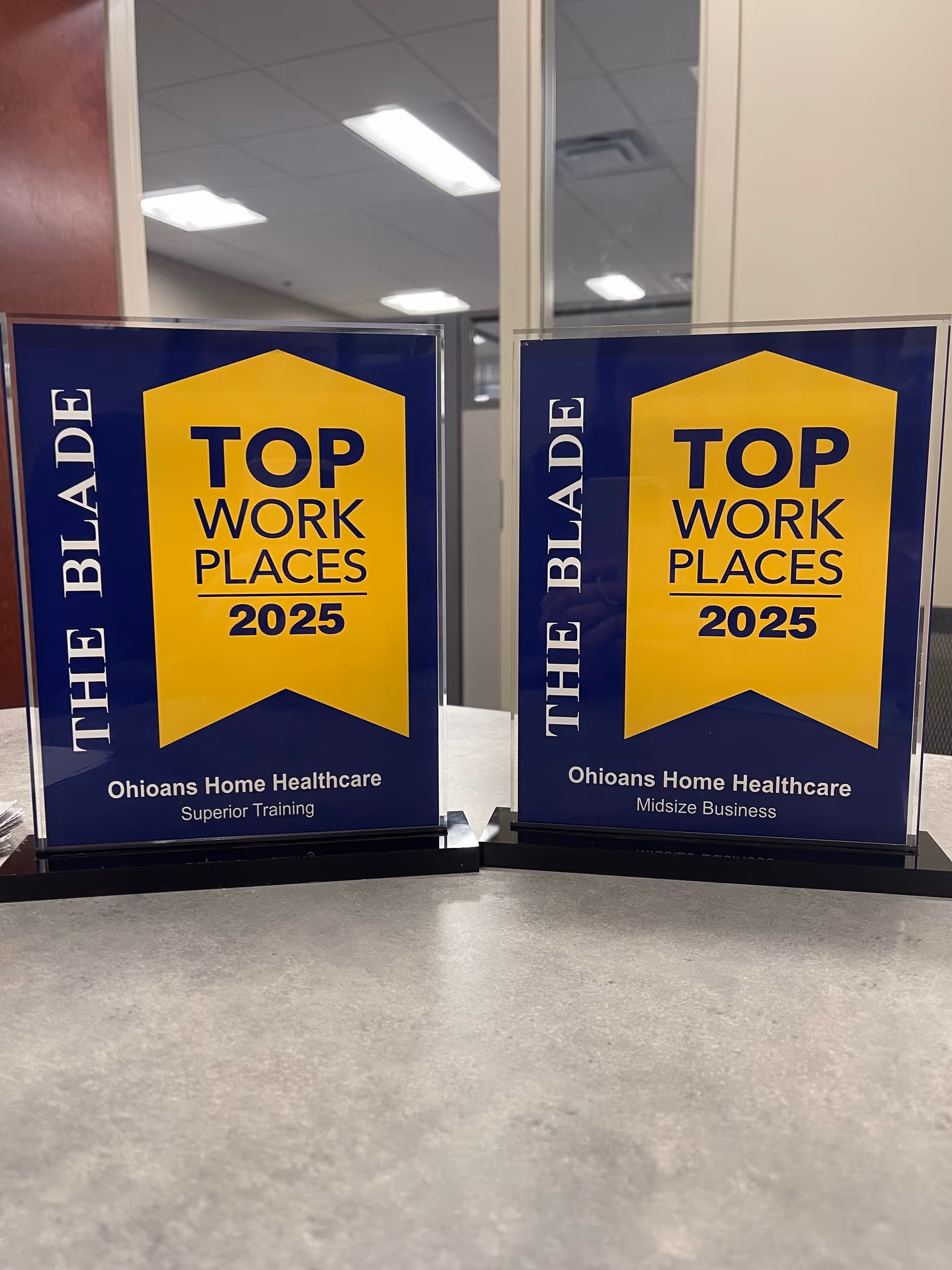 Two blue and yellow Top Work Places 2025 signs for Ohioans Home Healthcare on a desk.