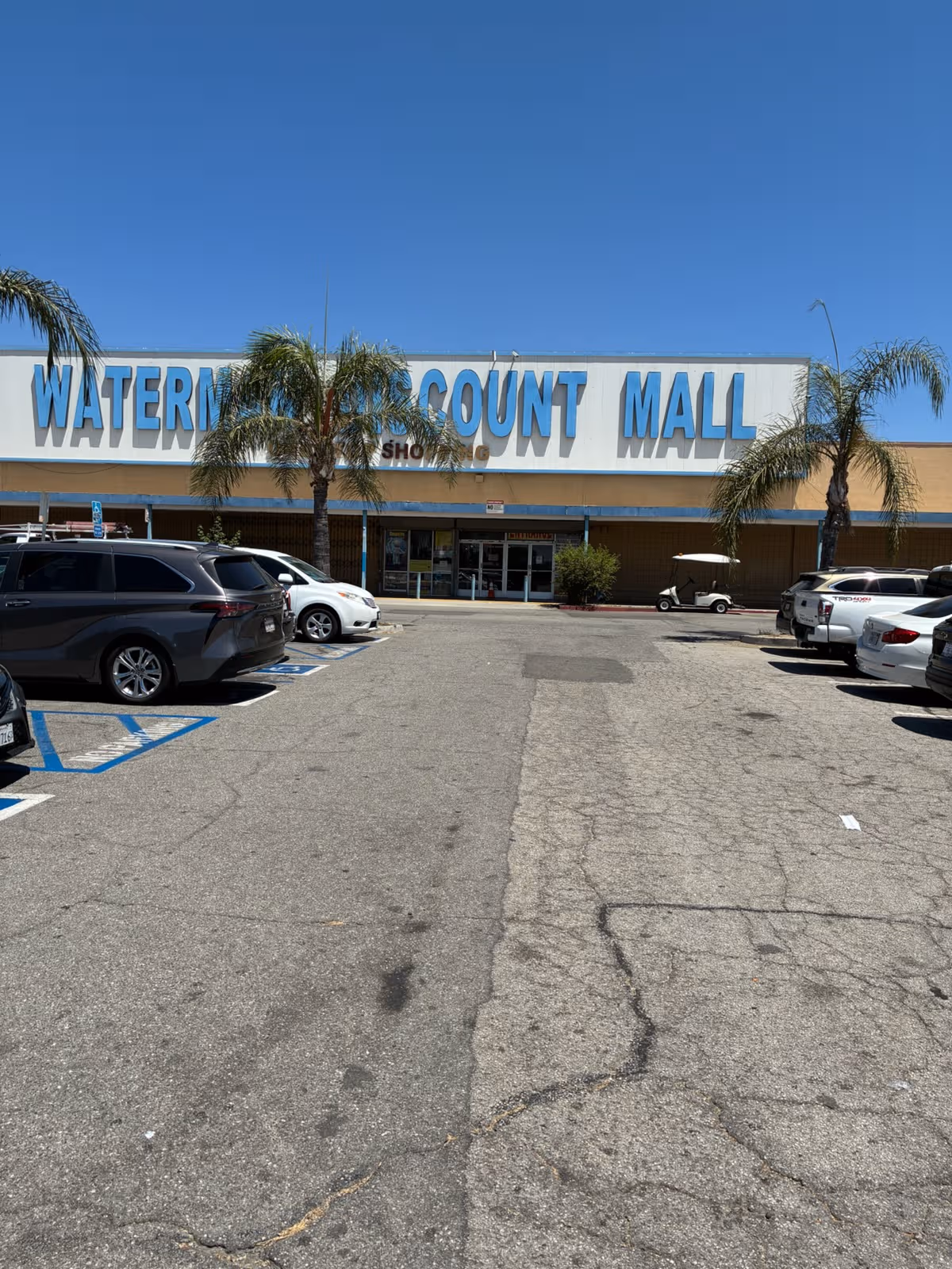 Exterior view of a large shopping mall facade with blue signage, palm trees, and parked cars.