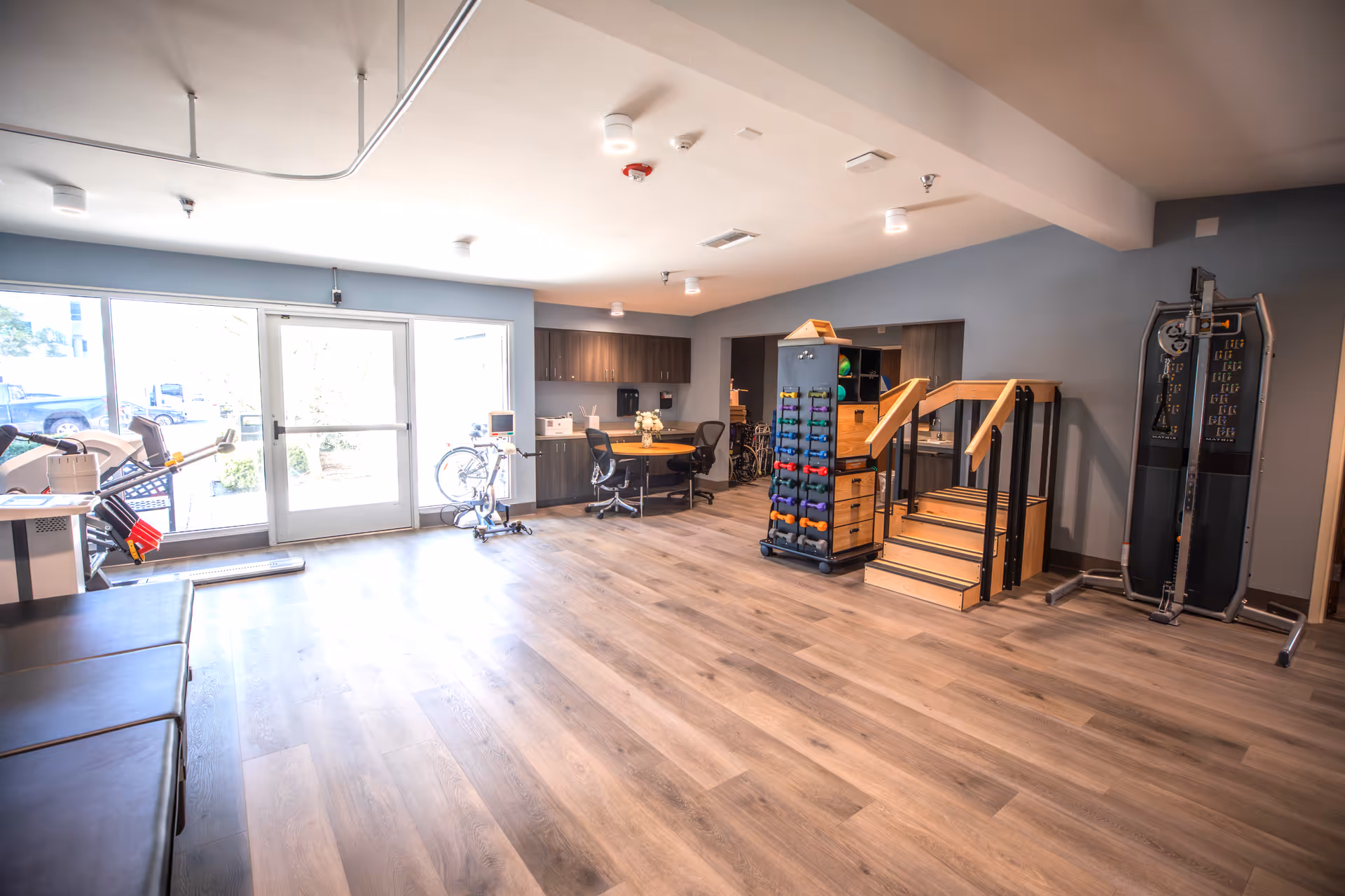 Bright indoor fitness area with weights, stairs, and cardio equipment near a large window.