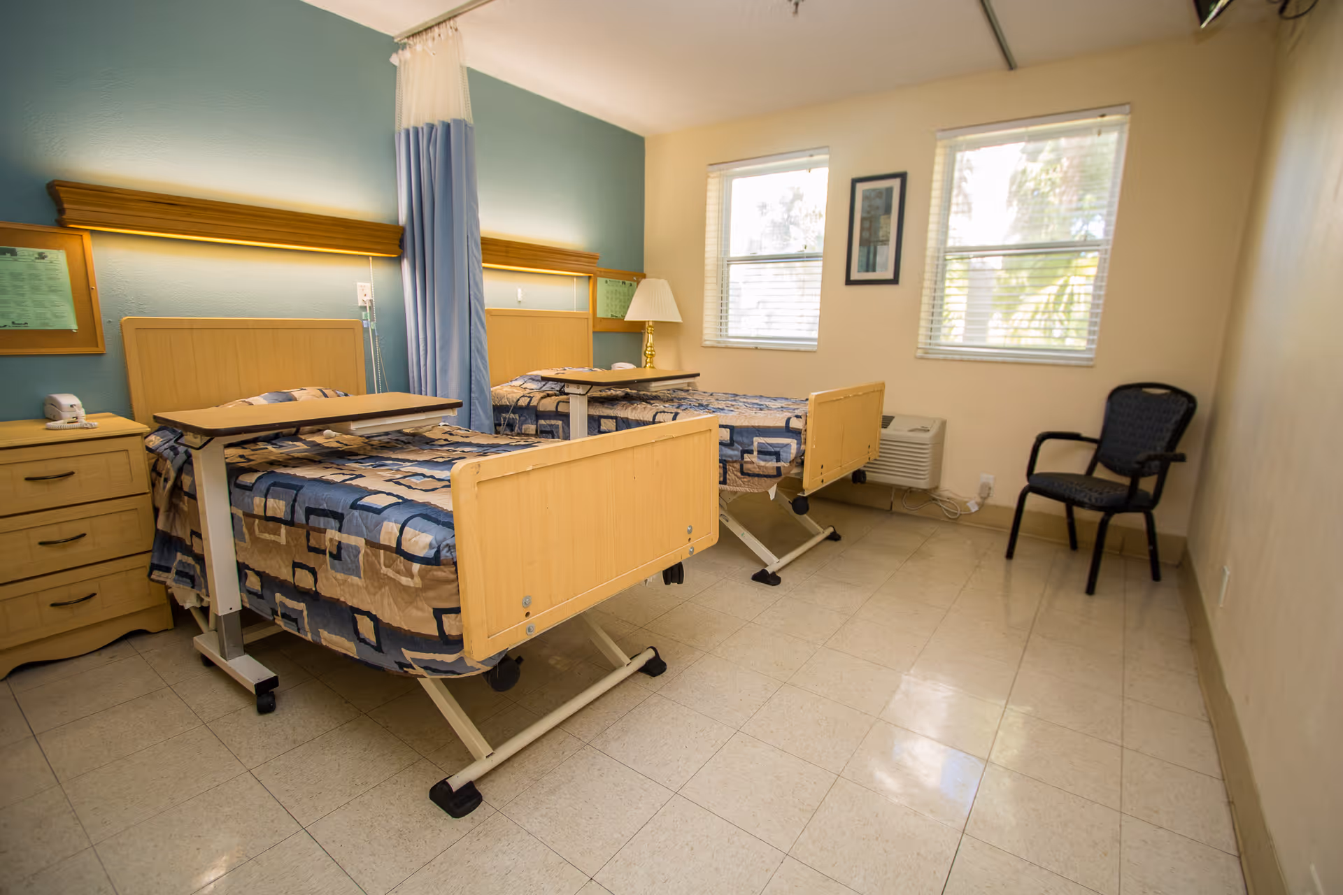 A shared bedroom in a care center with two hospital-style beds separated by a blue privacy curtain. Each bed has a patterned bedspread and an overbed table. There is a wooden nightstand with a telephone and a lamp between the beds. Two windows with blinds allow natural light into the room, and a single chair is placed against the wall.