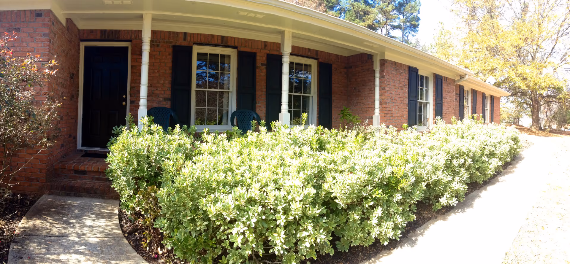Brick senior living facility front with a porch, chairs, and trimmed hedges.