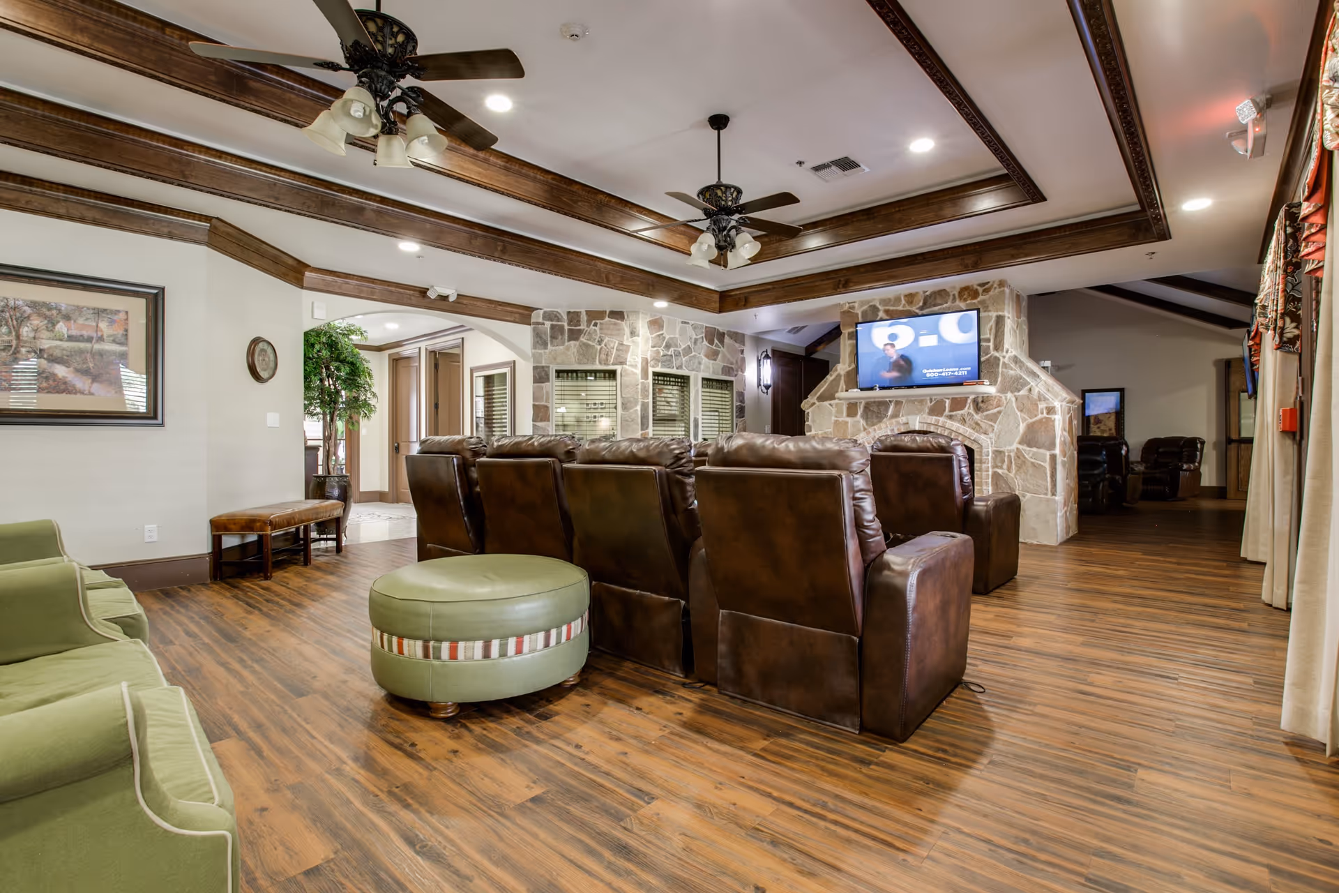 A spacious living room area in Avalon Memory Care featuring a row of brown leather recliners facing a stone fireplace with a mounted flat-screen TV. The room has wooden flooring, two ceiling fans with lights, green upholstered chairs, a round green ottoman, and decorative wooden ceiling beams. There is a framed picture on the wall and a potted plant near an archway leading to another room.