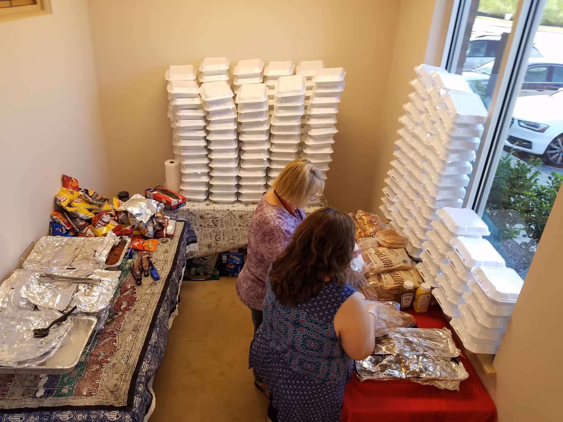 Two NuAngels staff pack meals at a prep table beside stacks of takeout containers.