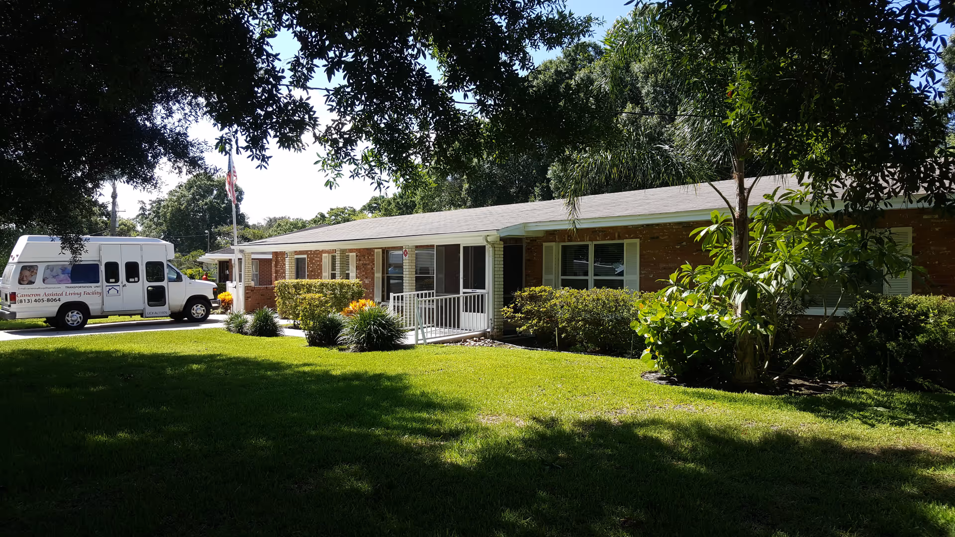 Front exterior of a single-story brick assisted living building with a green lawn, shrubs, and a white facility van parked by the driveway.