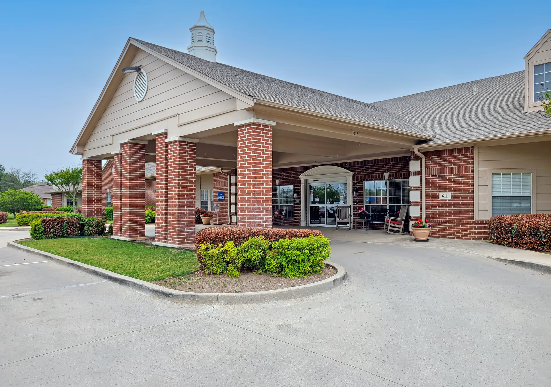 Exterior view of Morada Burleson facility entrance featuring a covered drop-off area with brick pillars, a paved driveway, green shrubs, and a clear blue sky.