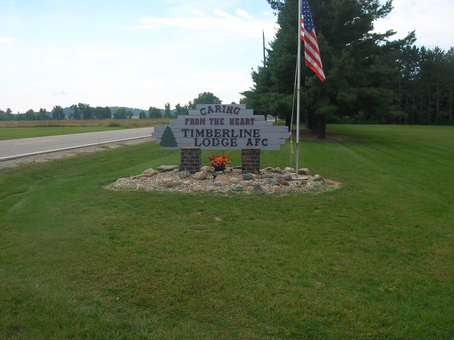 A roadside sign for Timberline Lodge AFC with the phrase 'Caring from the Heart' displayed above the facility name. The sign is mounted on two brick pillars surrounded by rocks and flowers, with an American flag on a flagpole to the right. The background shows a grassy area, trees, and a road extending into the distance under a partly cloudy sky.