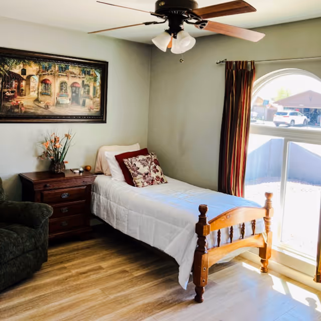 Cozy bedroom with a single wooden bed, nightstand, sofa, artwork, ceiling fan, and large window.