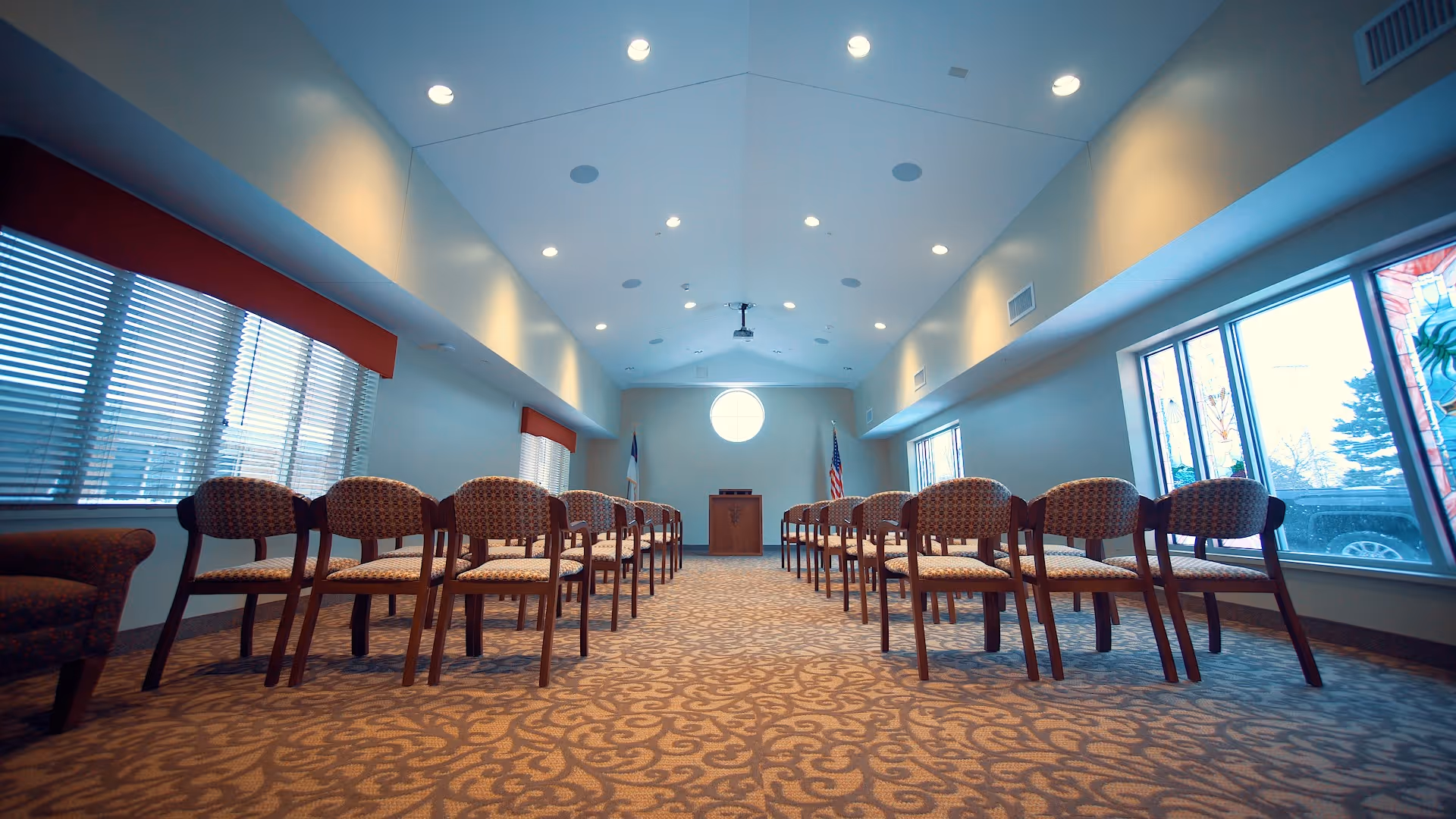 A spacious room with rows of patterned chairs arranged facing a wooden podium at the front. The room has large windows on both sides with blinds, a patterned carpet, and recessed ceiling lights. An American flag and another flag are positioned behind the podium.