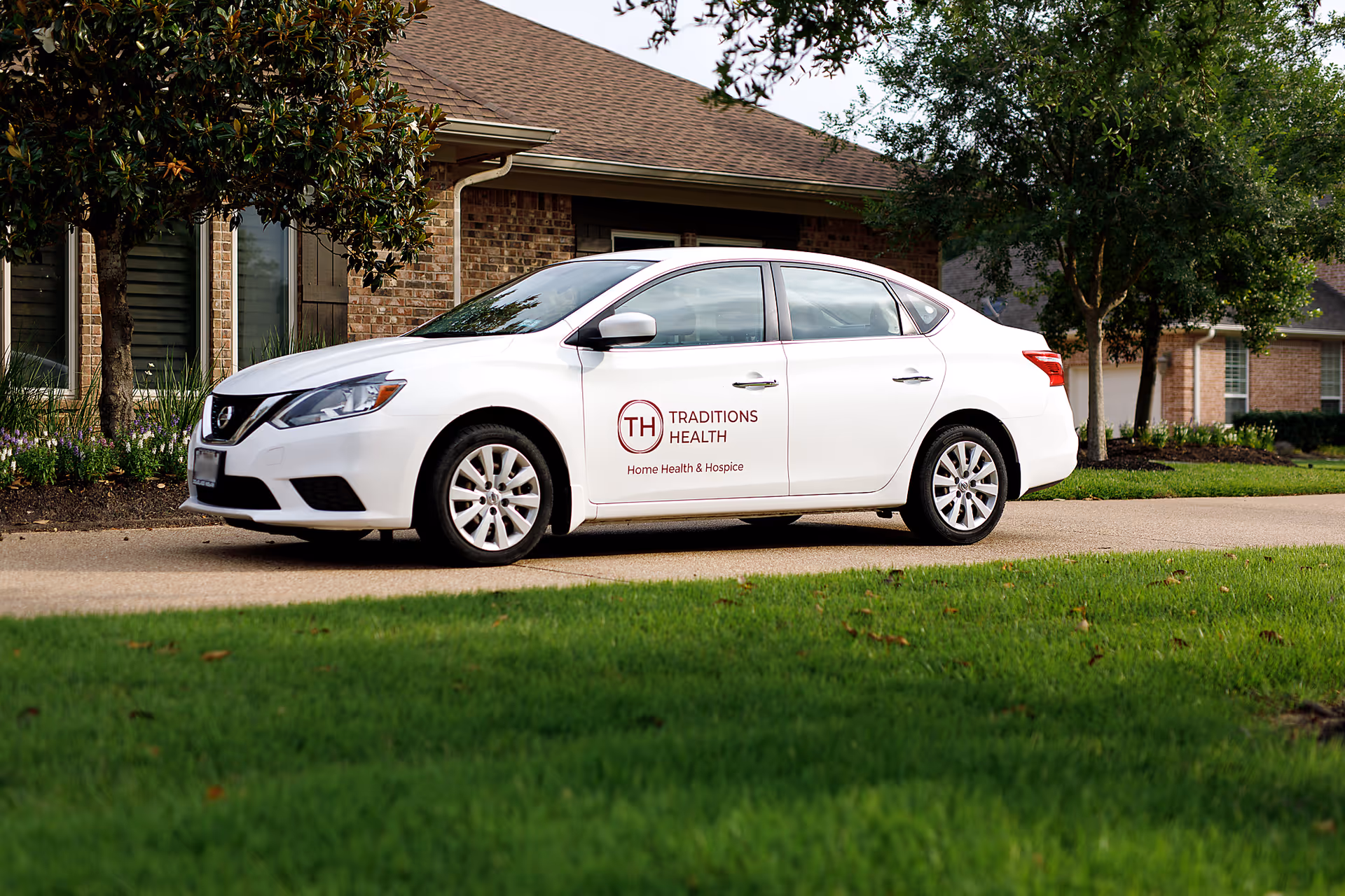 White car with Traditions Health logo parked in front of a brick senior living facility.