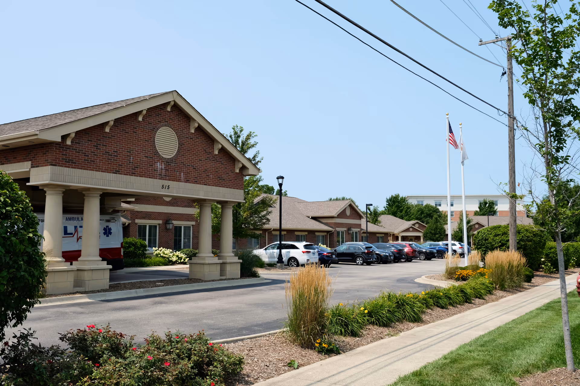 Front exterior of a brick memory care building with a covered columned entrance, parked cars, flagpoles, and landscaped grounds.