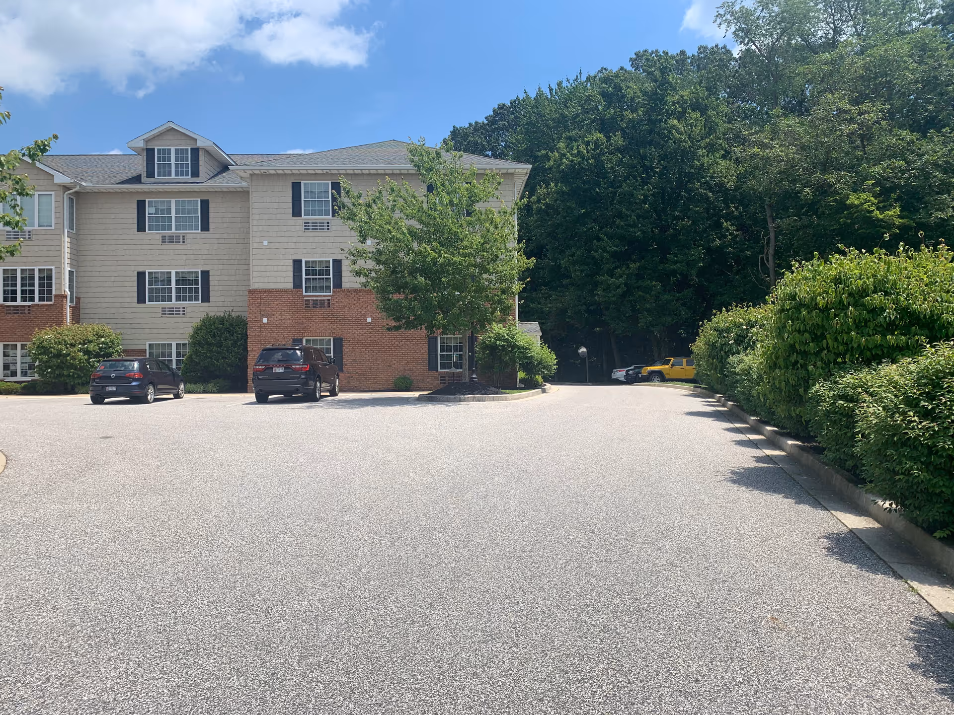 Three-story beige senior living building with brick accents, parking lot, and trees.