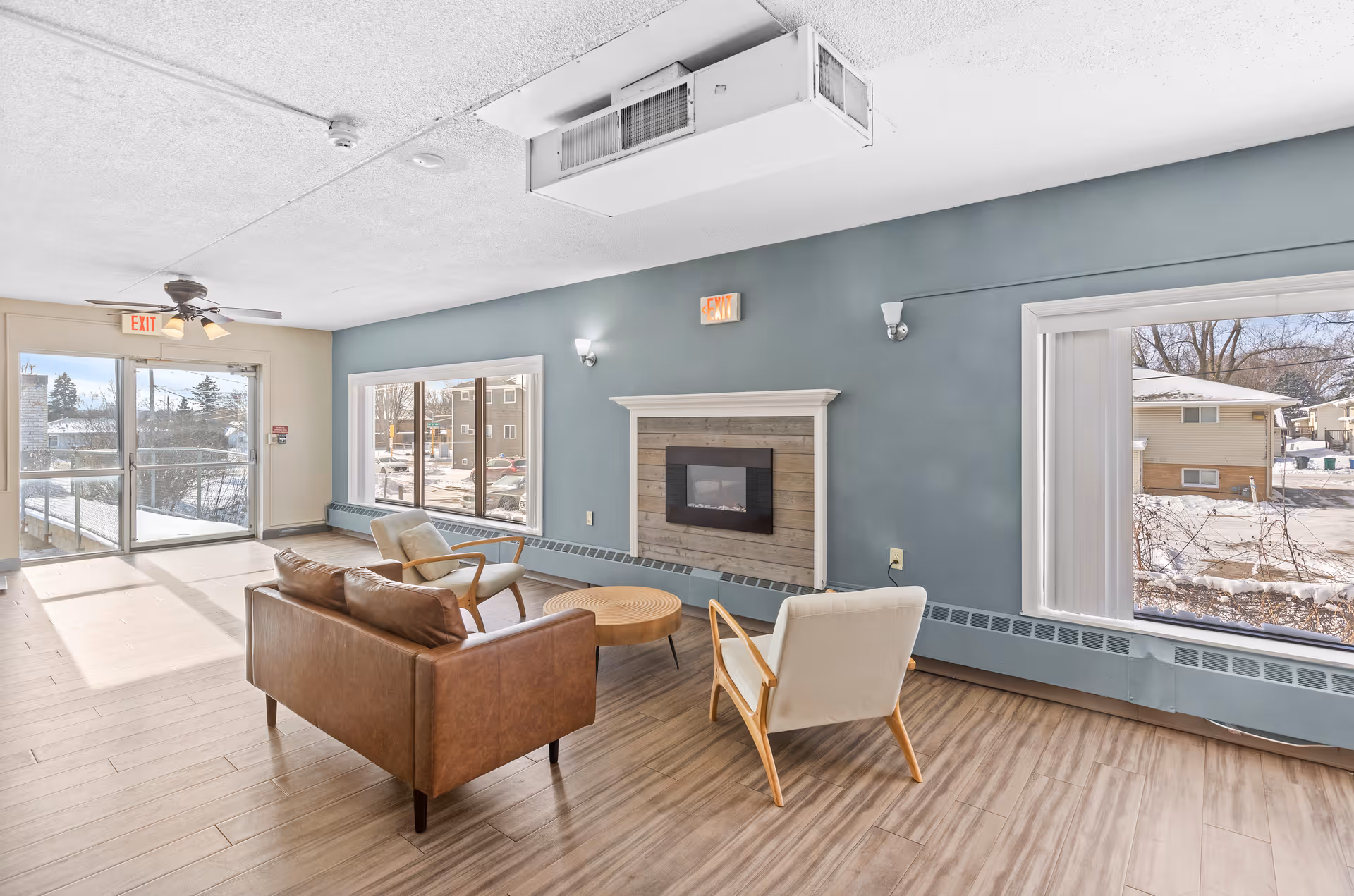 A bright and spacious living room area with large windows showing a snowy outdoor scene. The room features a brown leather sofa, two light-colored armchairs, a round wooden coffee table, a wall-mounted electric fireplace with a wooden frame, and a ceiling fan with lights. The walls are painted blue and beige, and the floor has wood-like tiles. There are exit signs above the doors and windows.