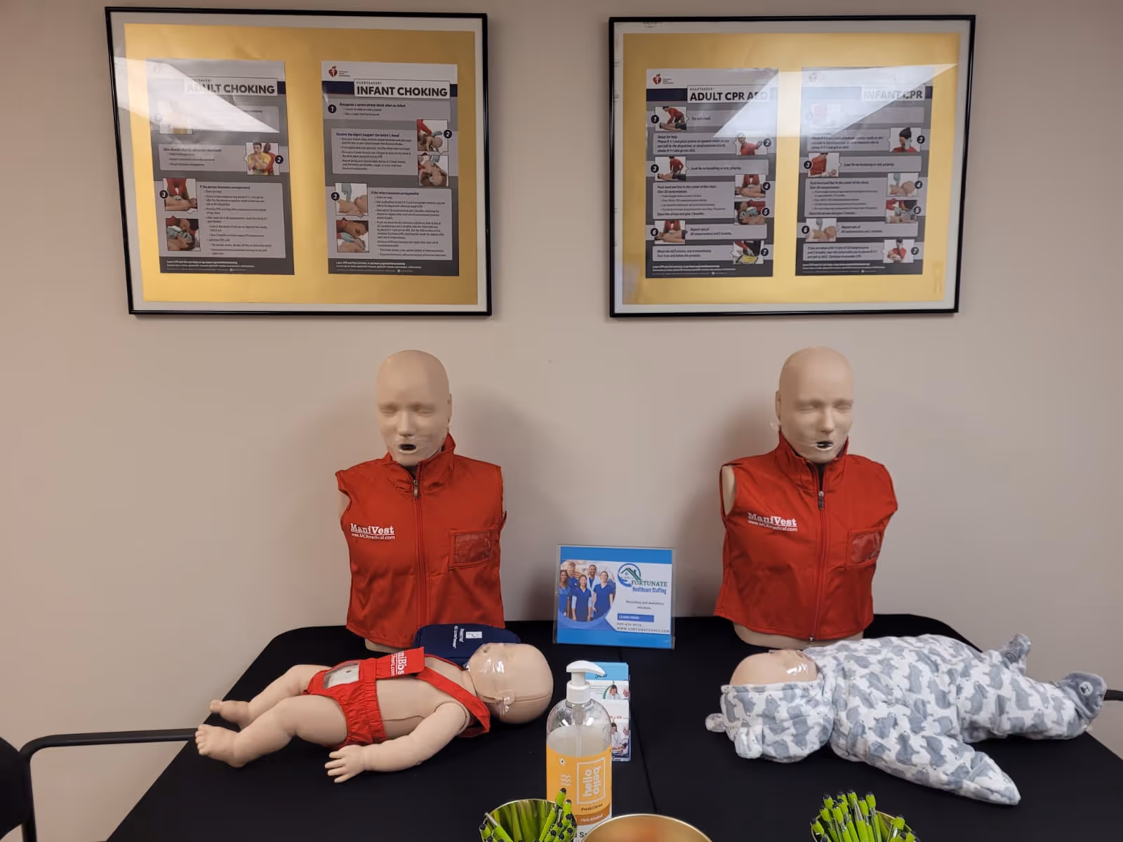 Two CPR mannequins in red vests with an infant dummy and posters on the wall.