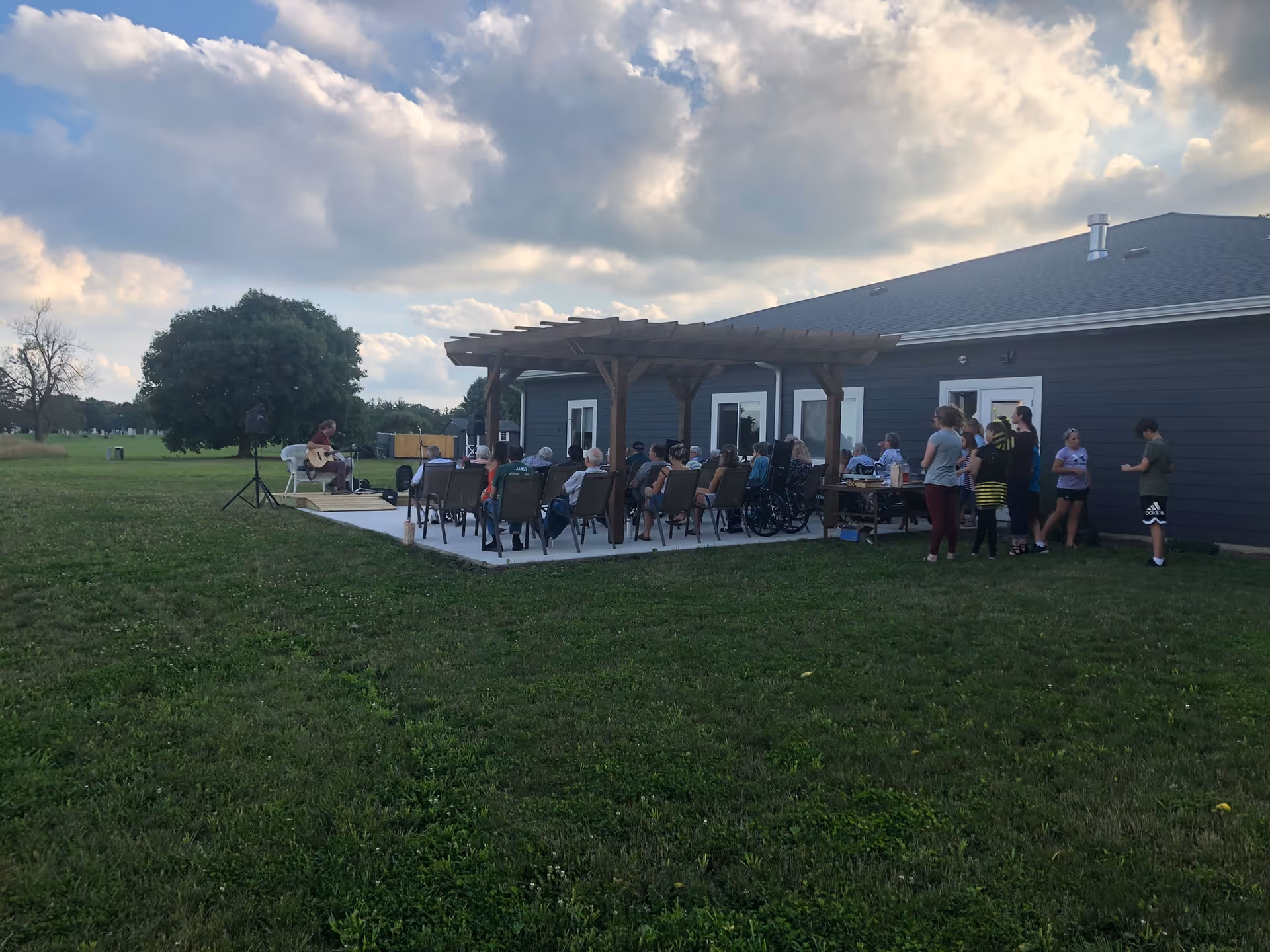 Residents and visitors seated on a patio under a wooden pergola outside a single-story building watching a musician perform.