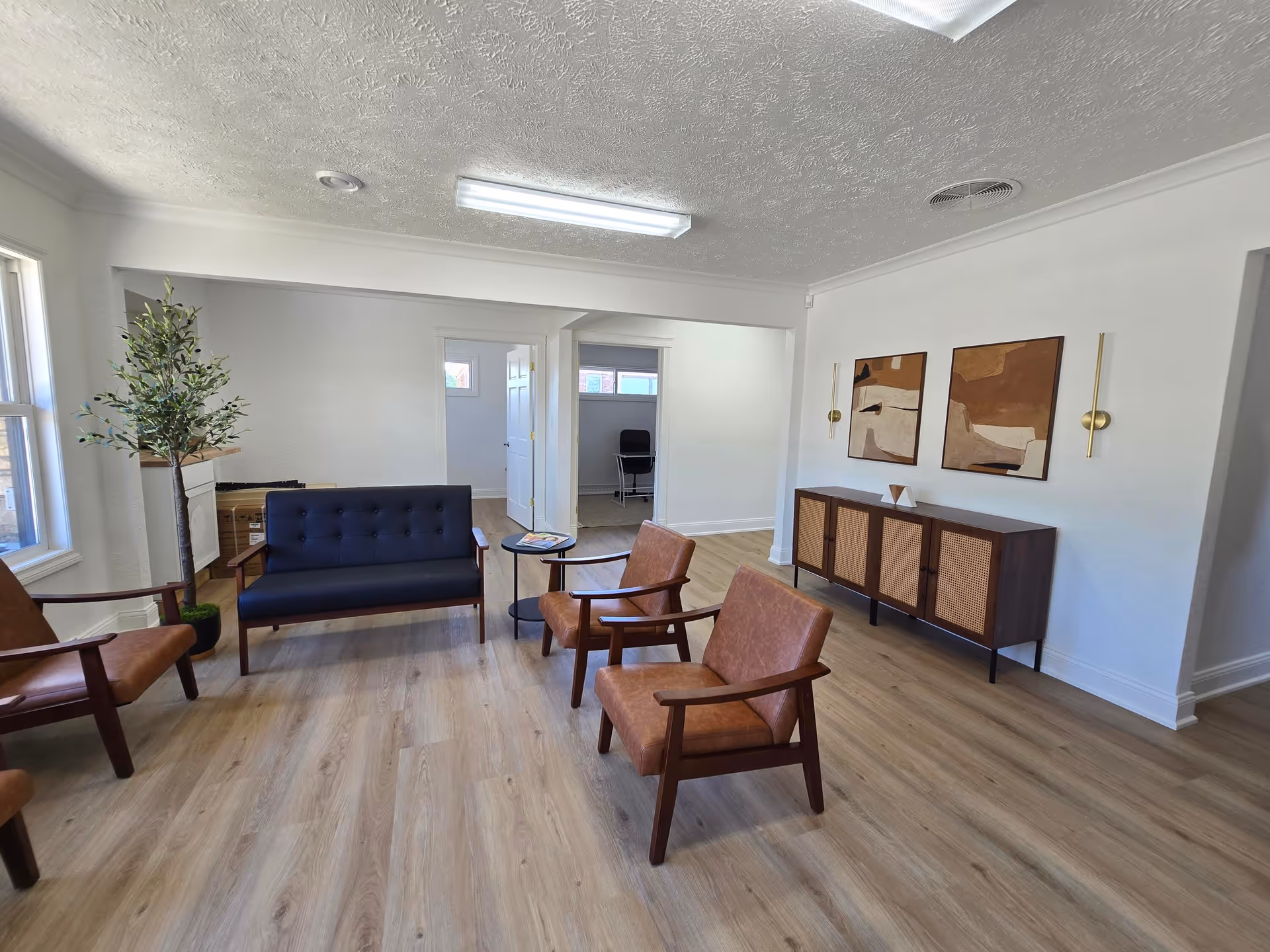 Bright living room with midcentury chairs, a dark sofa, and wall art in a senior facility.