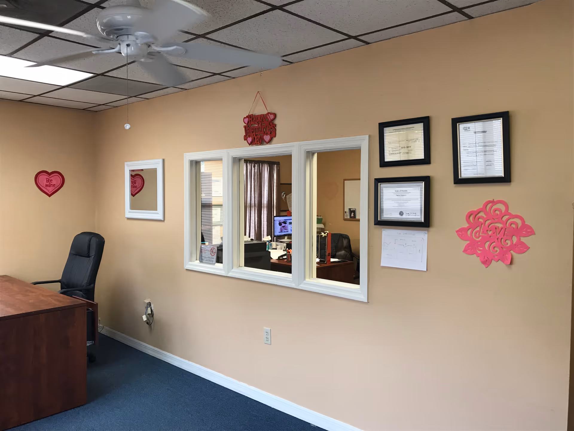 Beige office room with desk, chair, pass-through window, and pink wall decor.