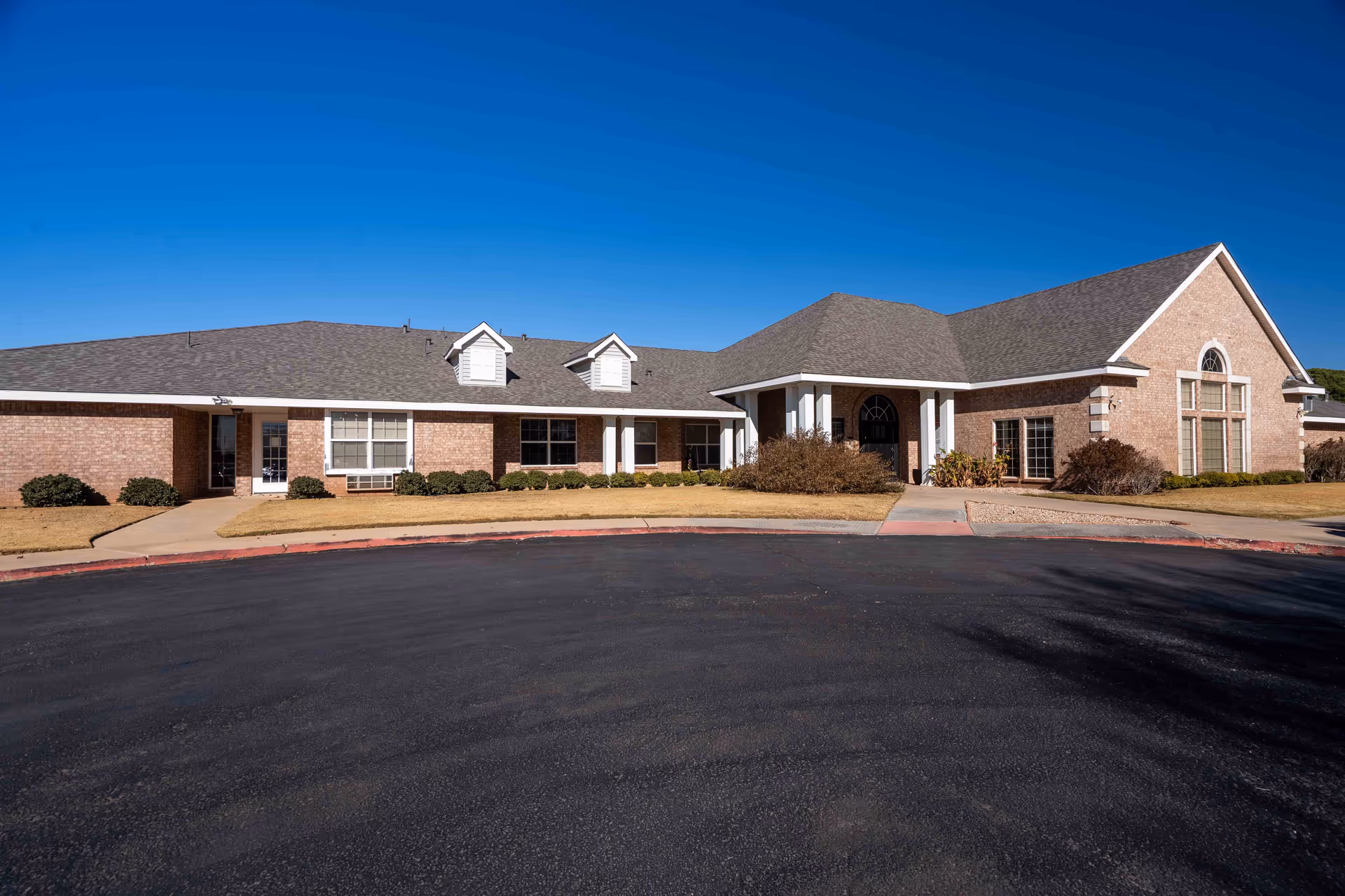 Brick senior living facility facade with wide entrance and manicured shrubs under a blue sky.