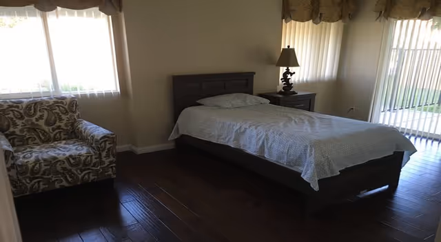 Bedroom in a senior living facility with bed, nightstands, lamp, and patterned chair.