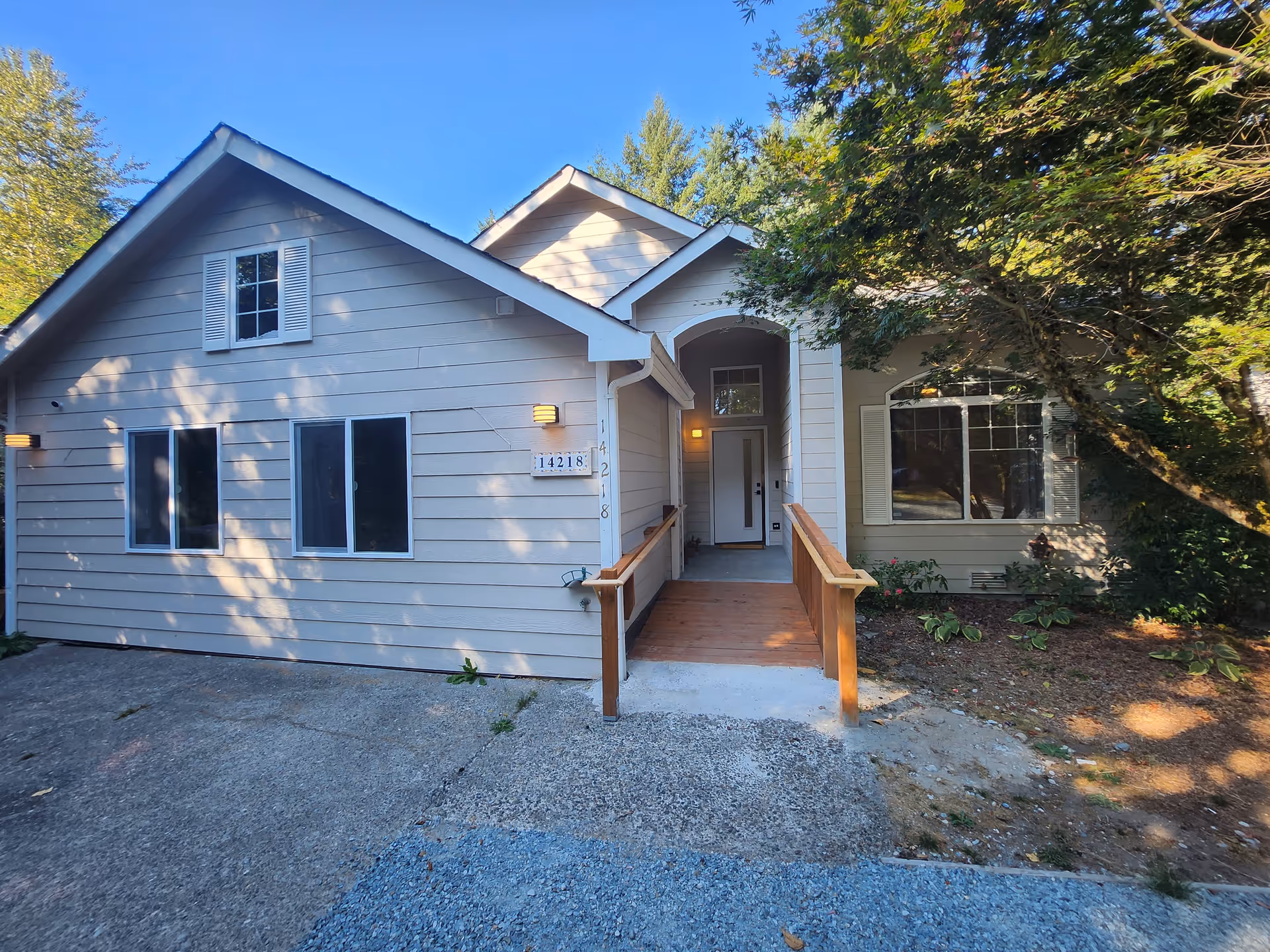 Front exterior of a beige single-story house with a covered entryway and wooden ramp, flanked by windows and trees.
