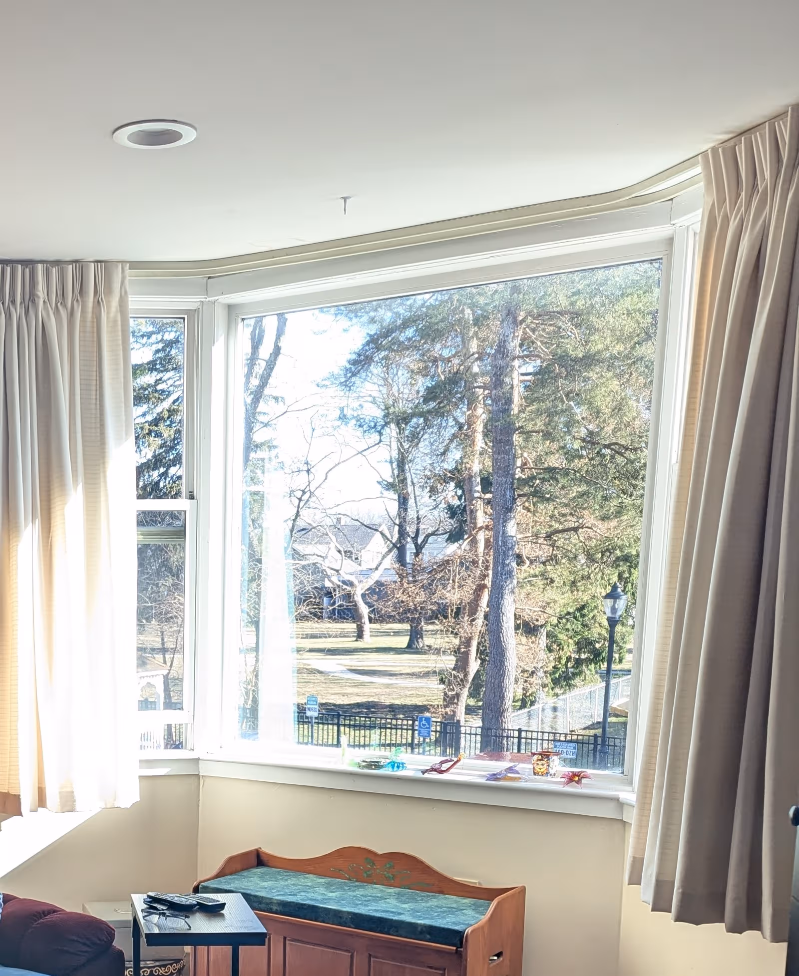 Bright interior corner with bay window, beige curtains, and a wooden bench by the window.