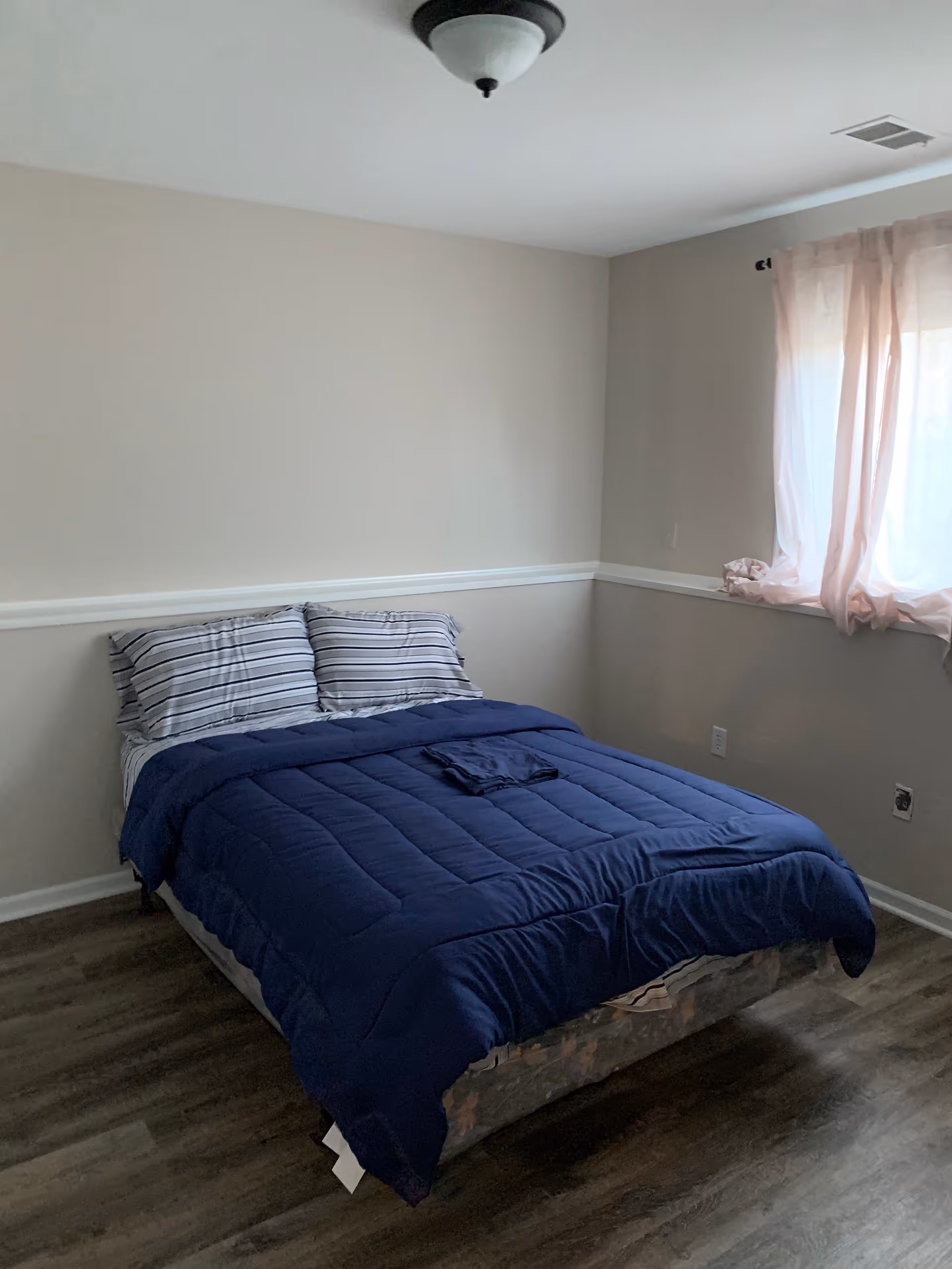 A small bedroom with a bed, blue comforter, striped pillows, and a window.