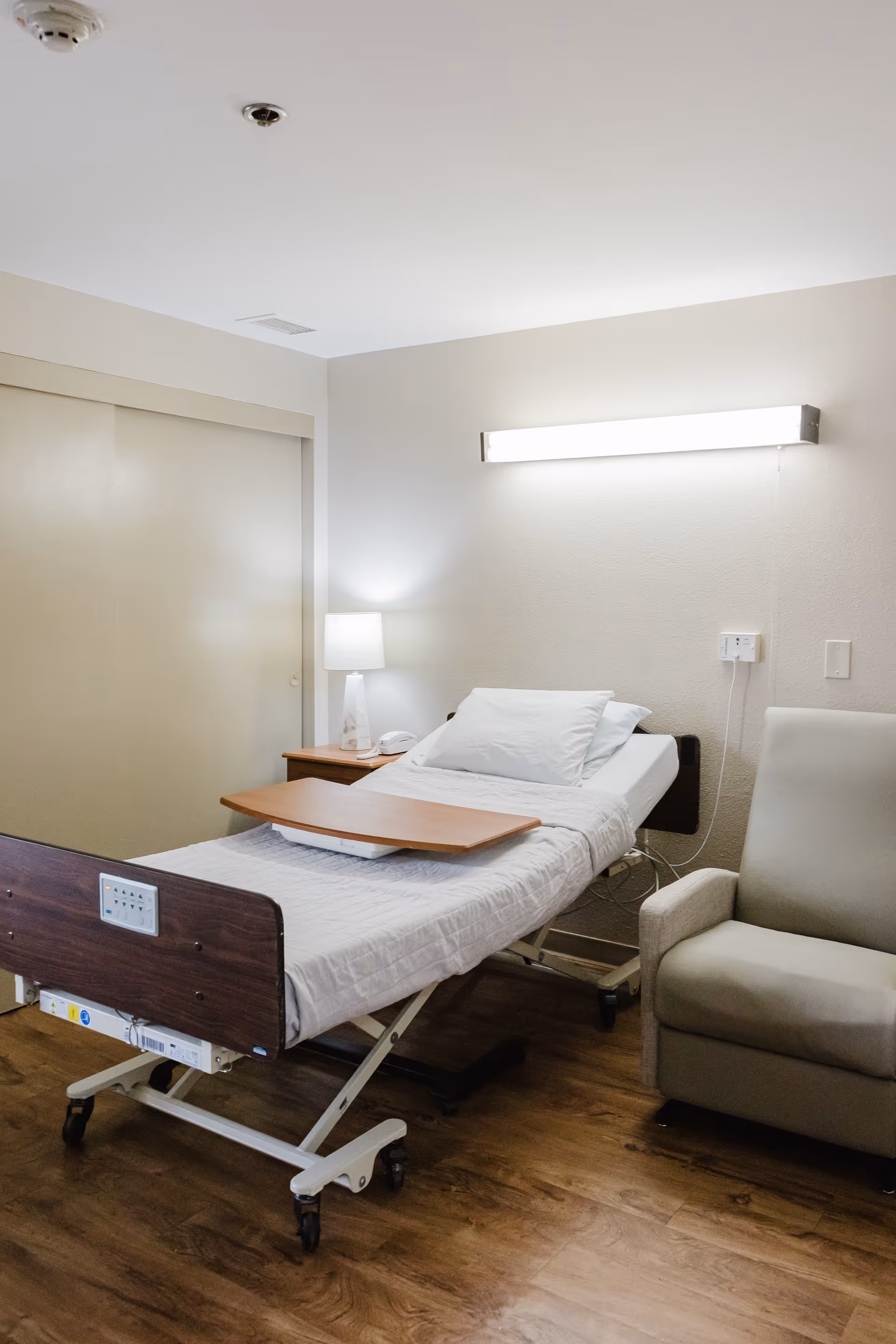 Hospital-style patient bedroom with adjustable bed, nightstand, lamp, and beige chair.