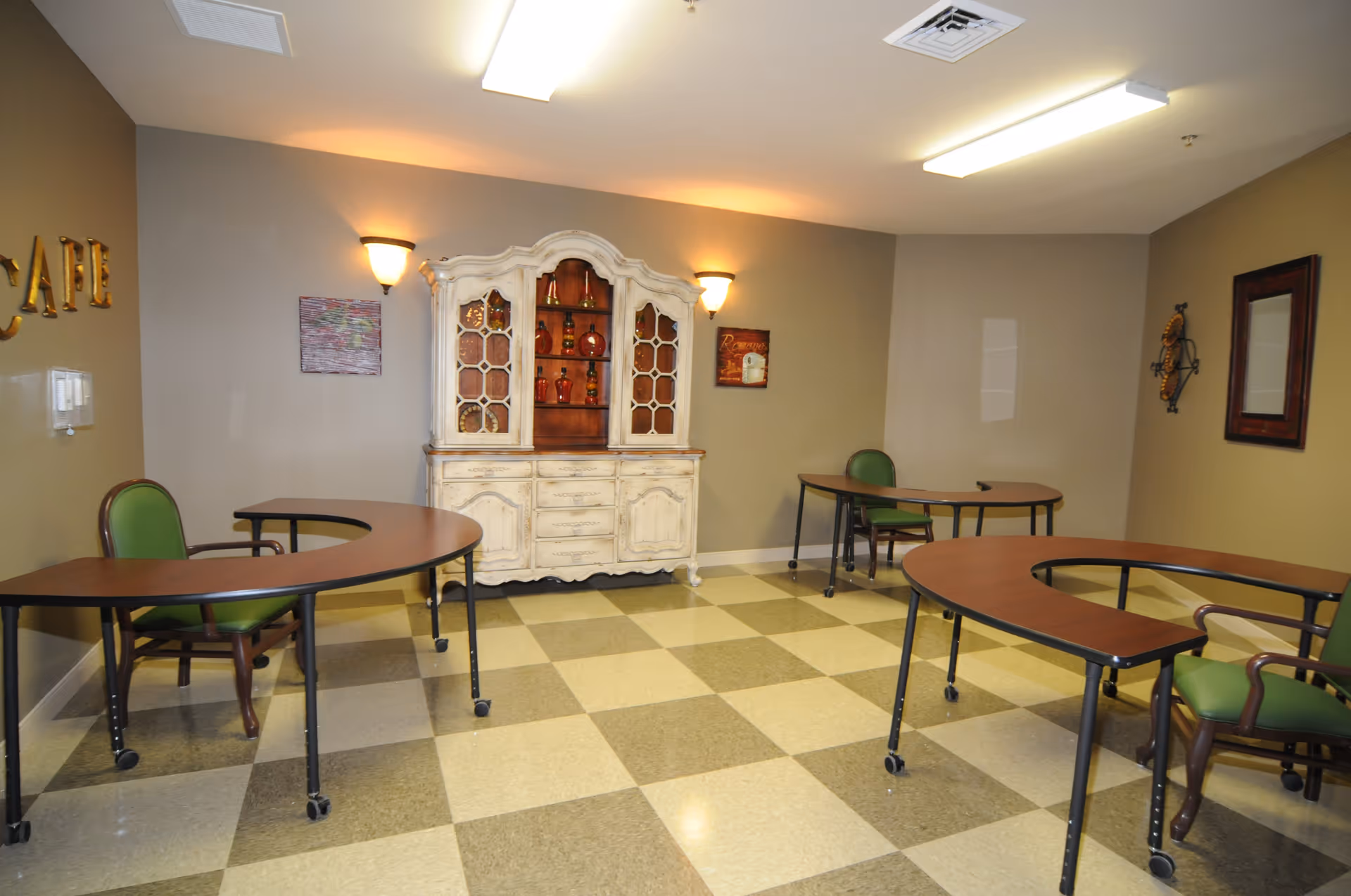 A small room with three curved tables on wheels, each paired with a green cushioned chair. The walls are painted beige, and there is a vintage white cabinet with glass doors displaying decorative items against the back wall. The floor has a checkered pattern with beige and light brown tiles. Wall sconces and ceiling lights illuminate the room.