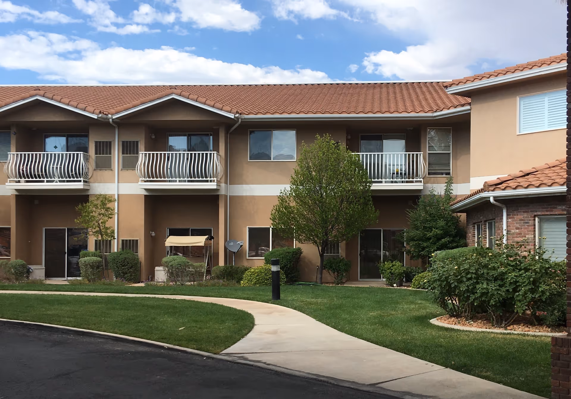 Exterior view of a two-story assisted living facility building with beige walls and red tile roof. The building features small balconies on the upper floor and sliding glass doors on the ground floor. There is a well-maintained lawn with bushes, trees, and a curved concrete walkway in front of the building under a partly cloudy sky.