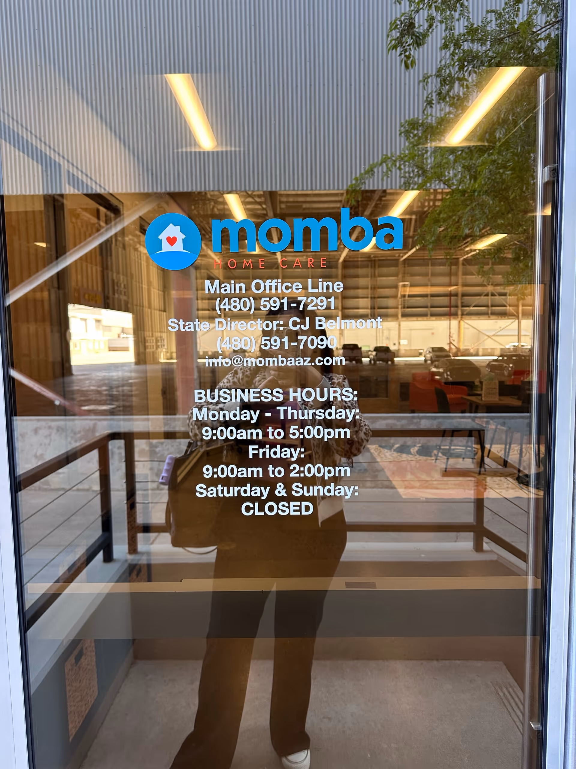 Glass storefront door shows Momba Home Care logo and business hours, with photographer's reflection.