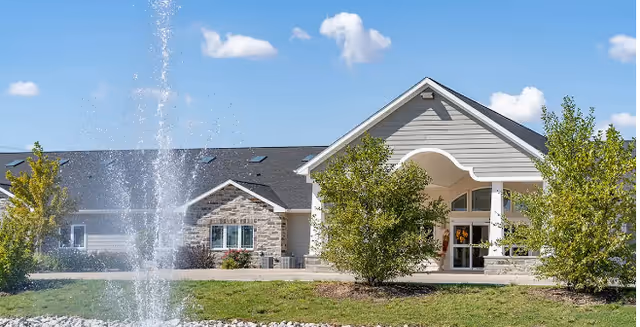Entrance facade of a single-story building with a fountain, landscaped shrubs, and a clear blue sky.