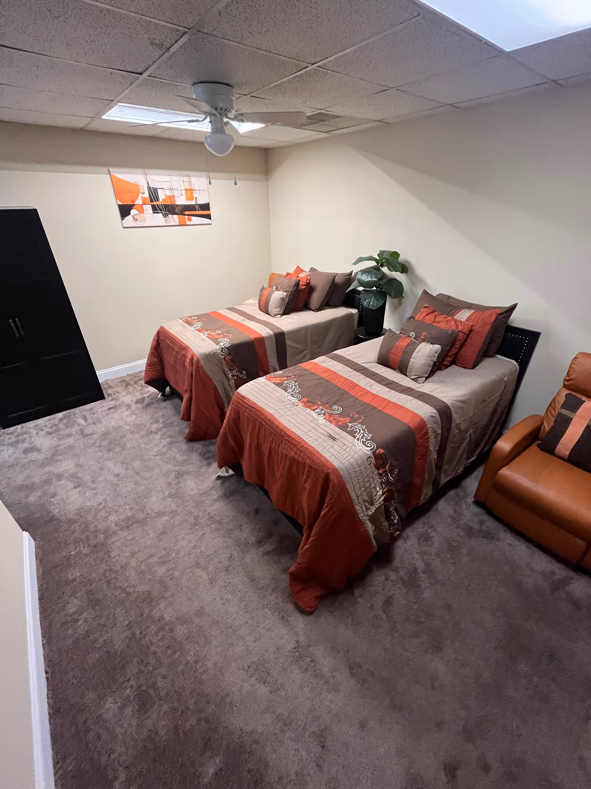 Two twin beds with red-brown bedding, cushions, a plant, chair, and wall art in a bedroom.
