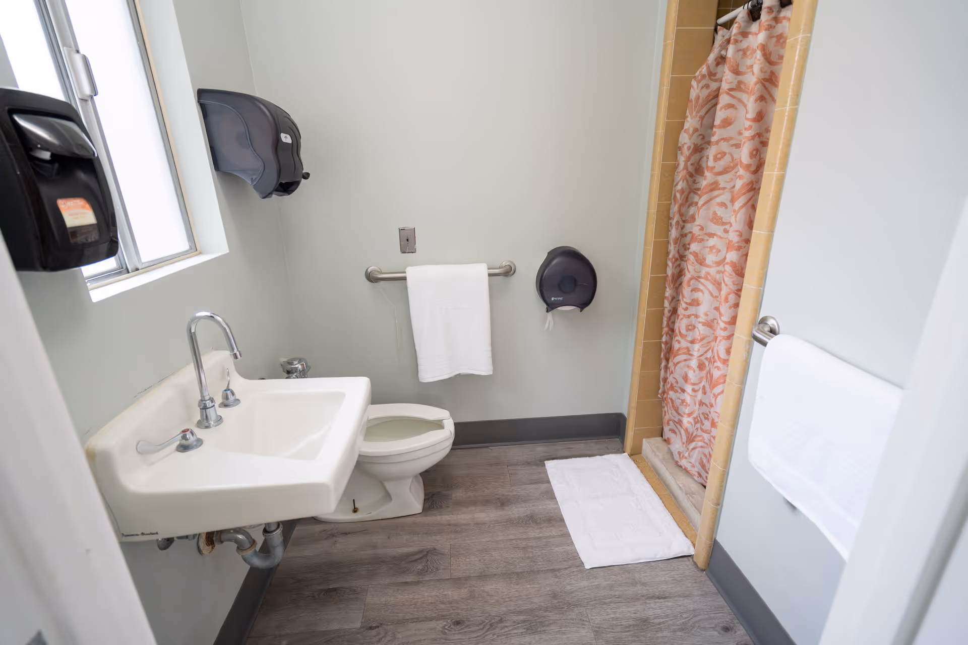 Compact bathroom with sink, toilet, shower, towel bars, and a window.