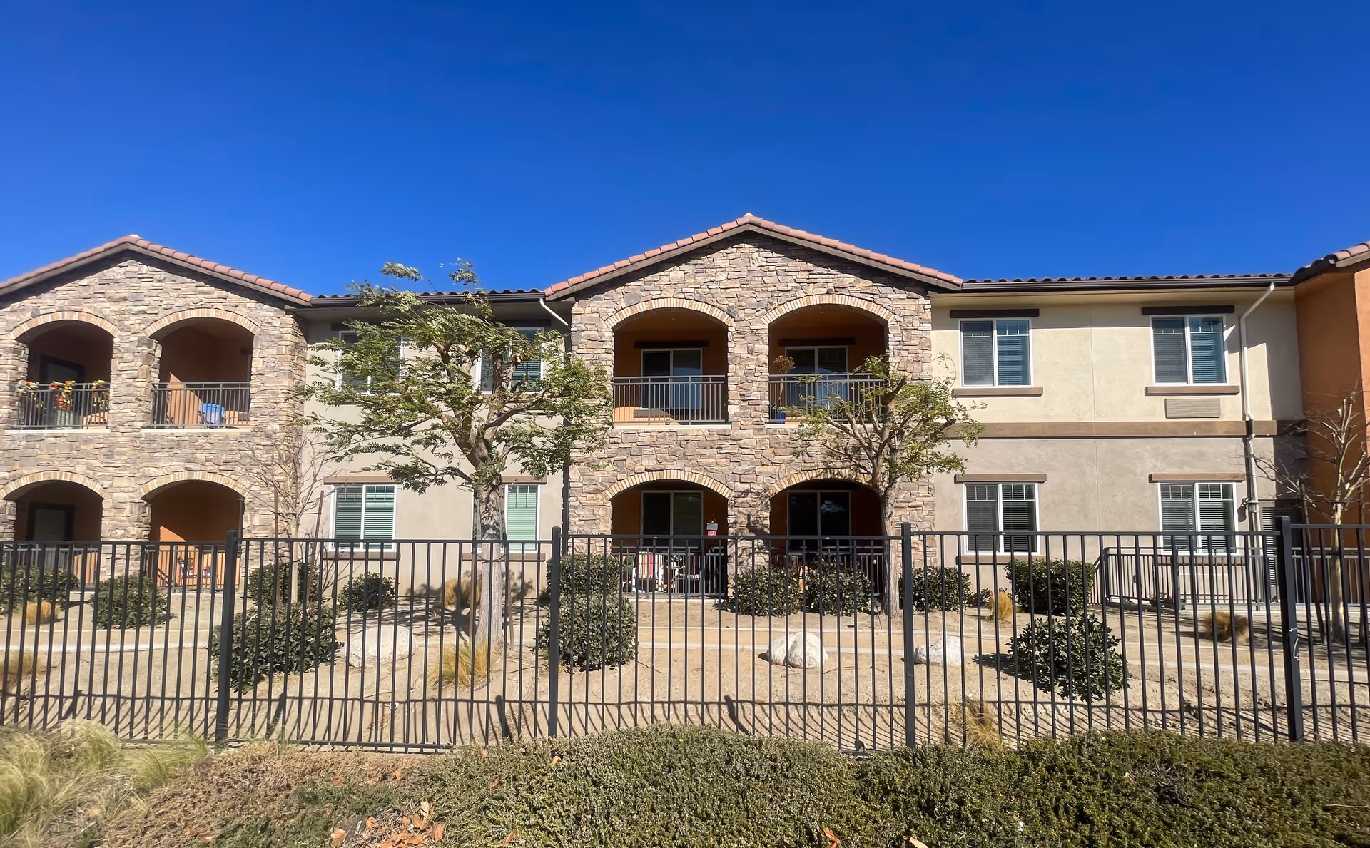 Exterior view of a two-story senior living facility building with stone and stucco facade, arched balconies, several windows, small trees, bushes, and a black metal fence in front under a clear blue sky.