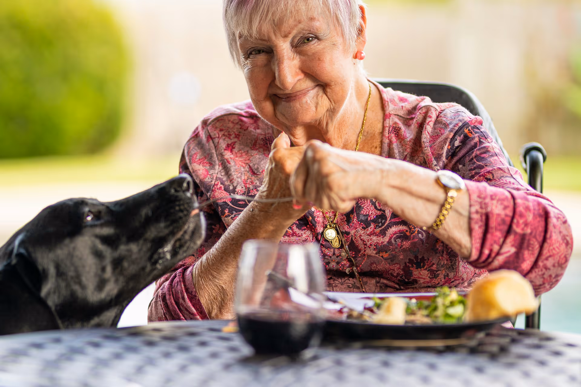 Senior woman outdoors at a dining table with a black dog beside her.